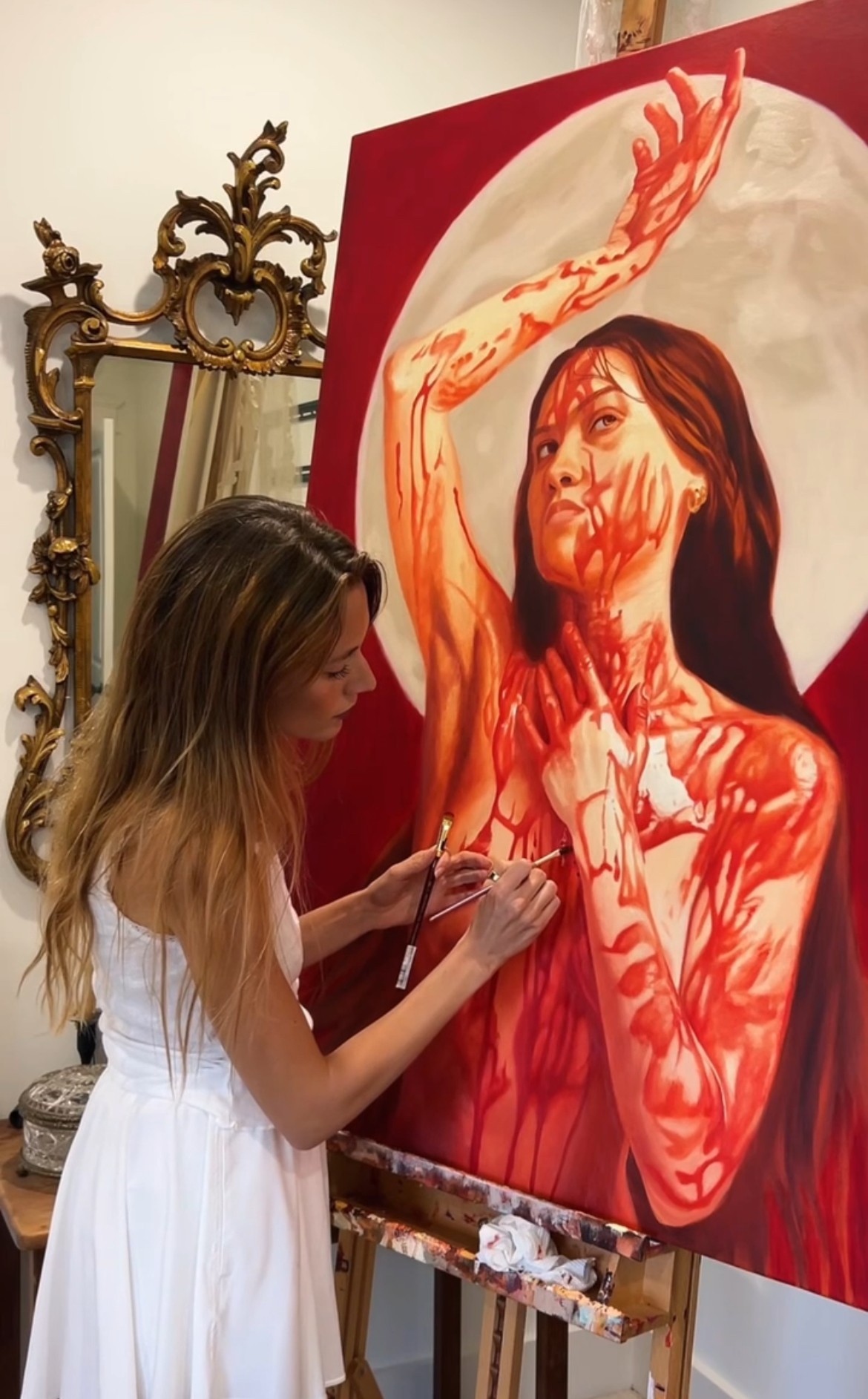 Woman painting a large portrait of a woman with long dark hair and red markings on her face and body, in an ornate room.