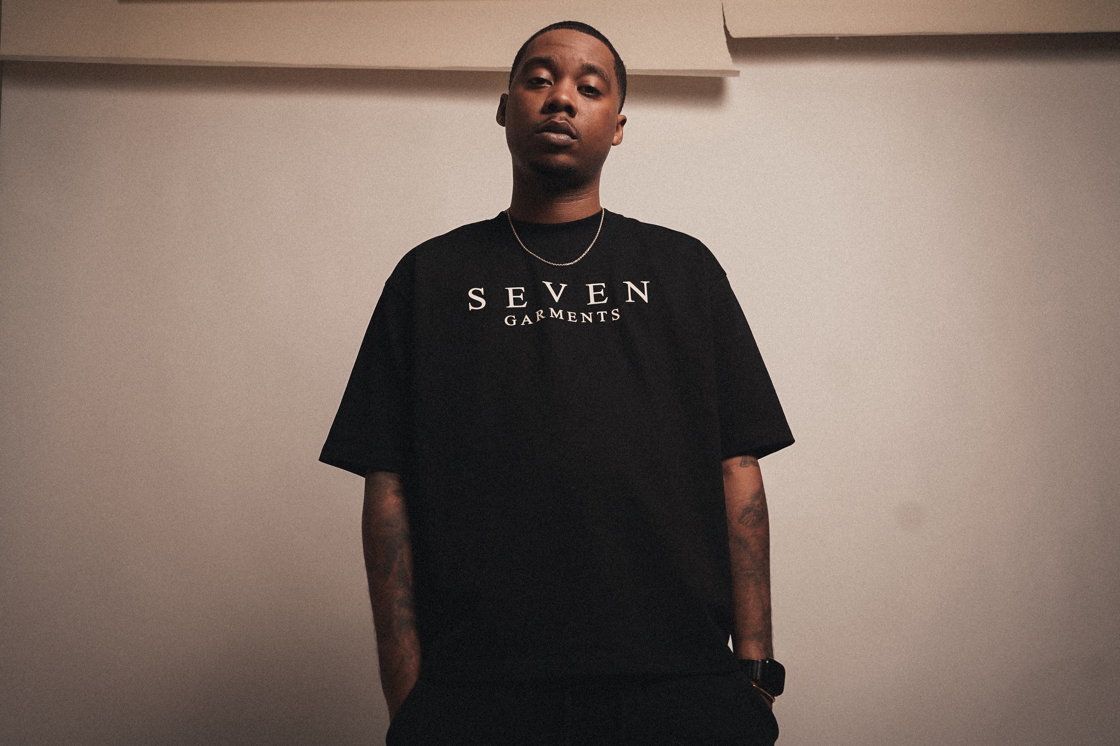 Young person with short hair wearing a black T-shirt with white text, standing against a plain wall.