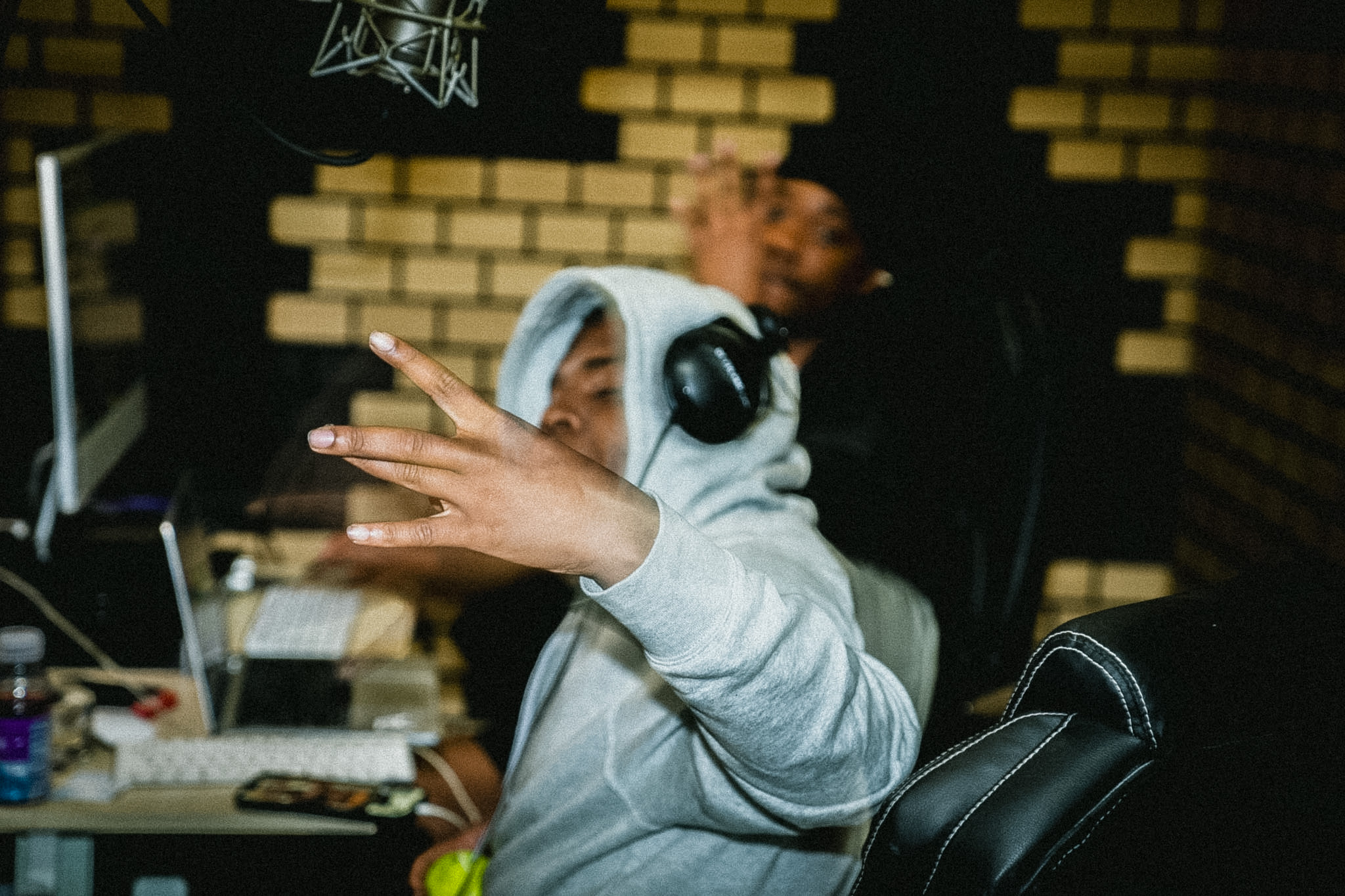 Person wearing a hoodie and headphones, making a peace sign with fingers, in a recording studio with brick wall background.