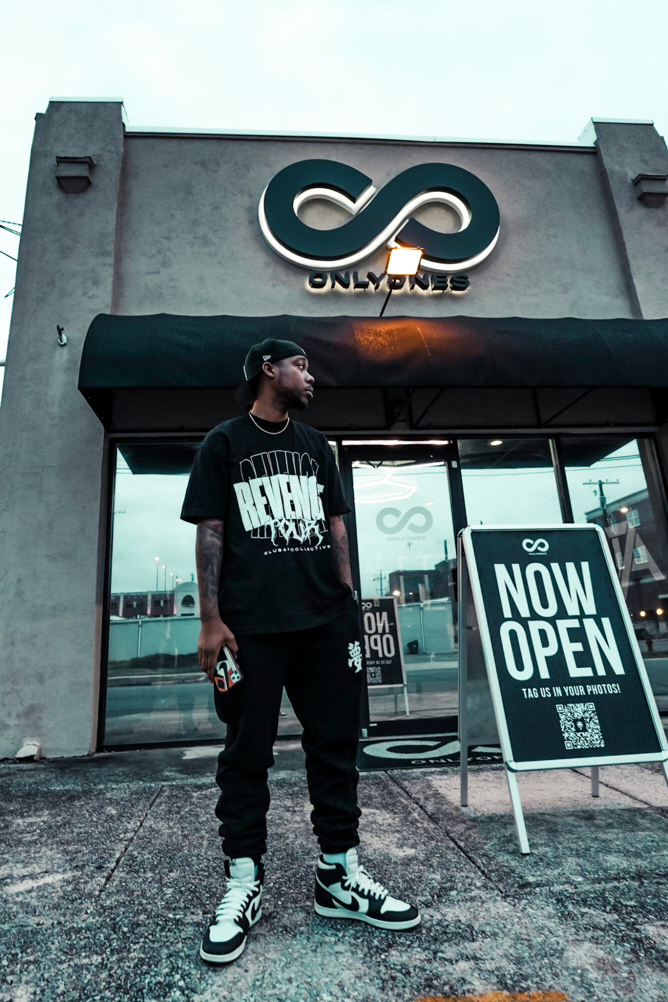 Young man standing outside a store with an infinity symbol logo and a 'Now Open' sign, cloudy sky background.