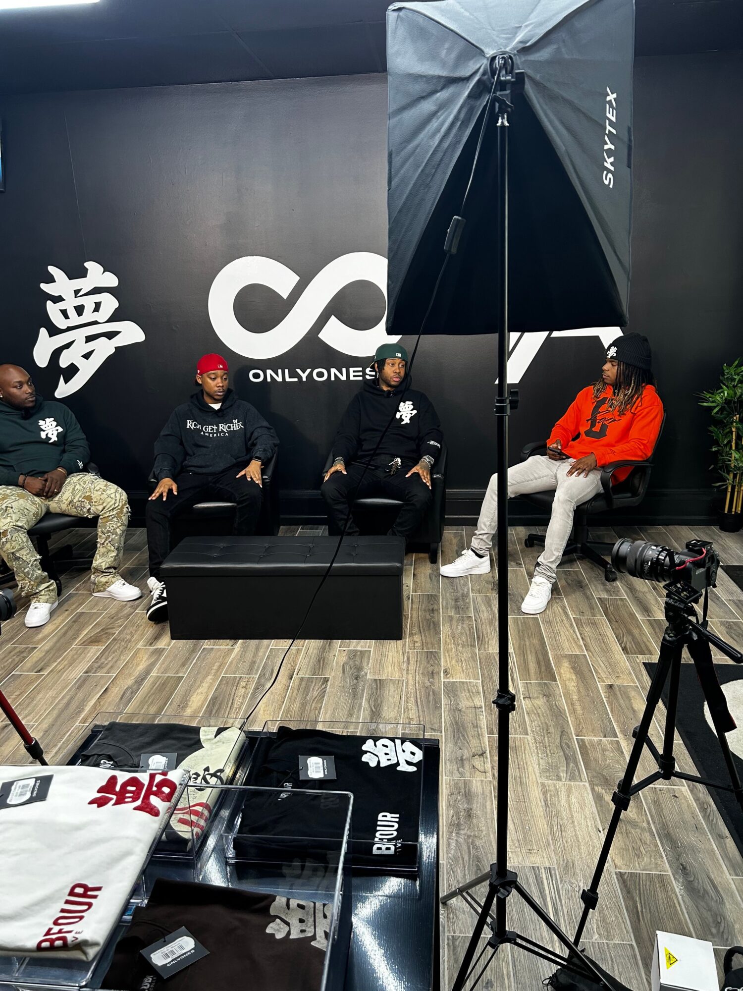 Three people sitting in a studio with a black background and a large white logo, surrounded by photography equipment.