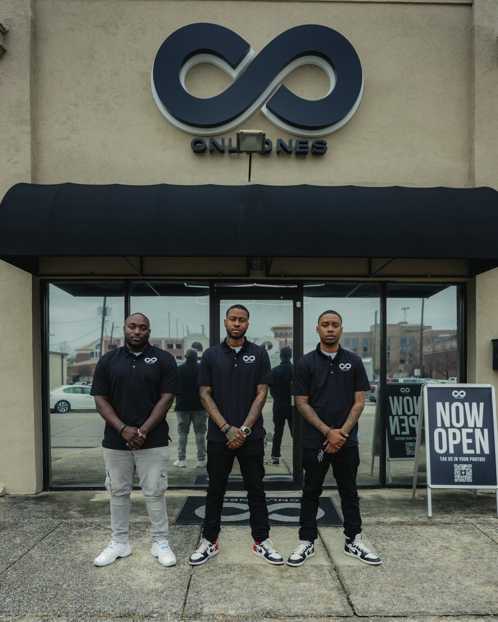 Three men stand outside a building with a large glass window and a sign that says 'NOW OPEN'.
