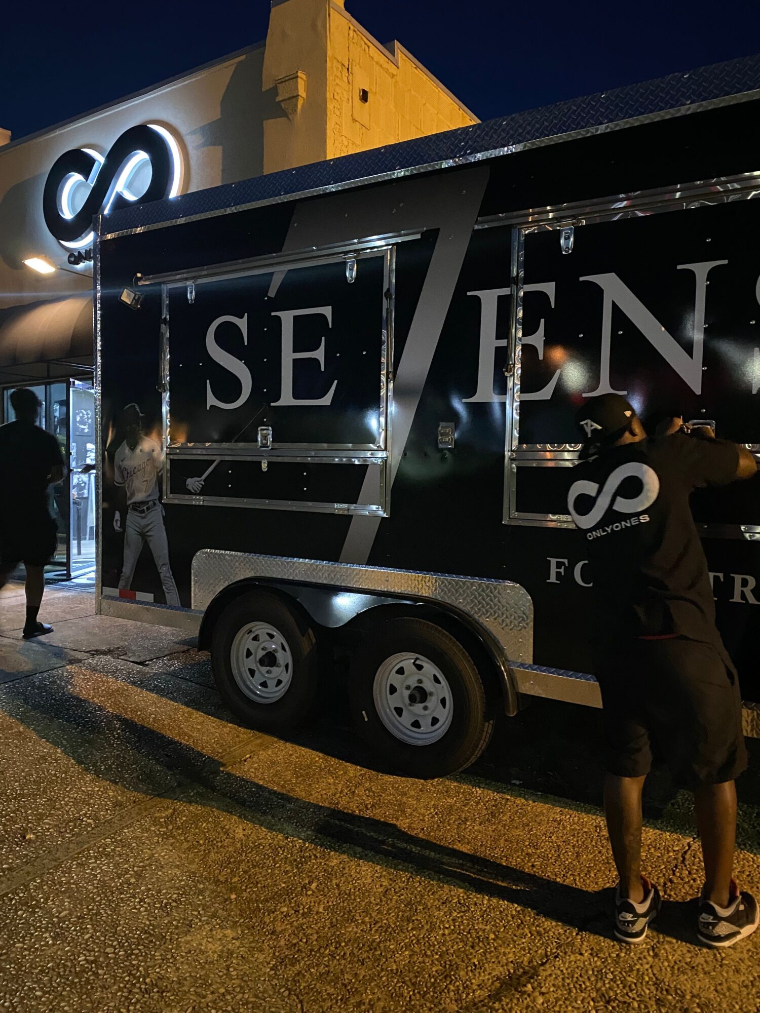Person standing next to a trailer with large text, on a paved surface at night, illuminated by lights.