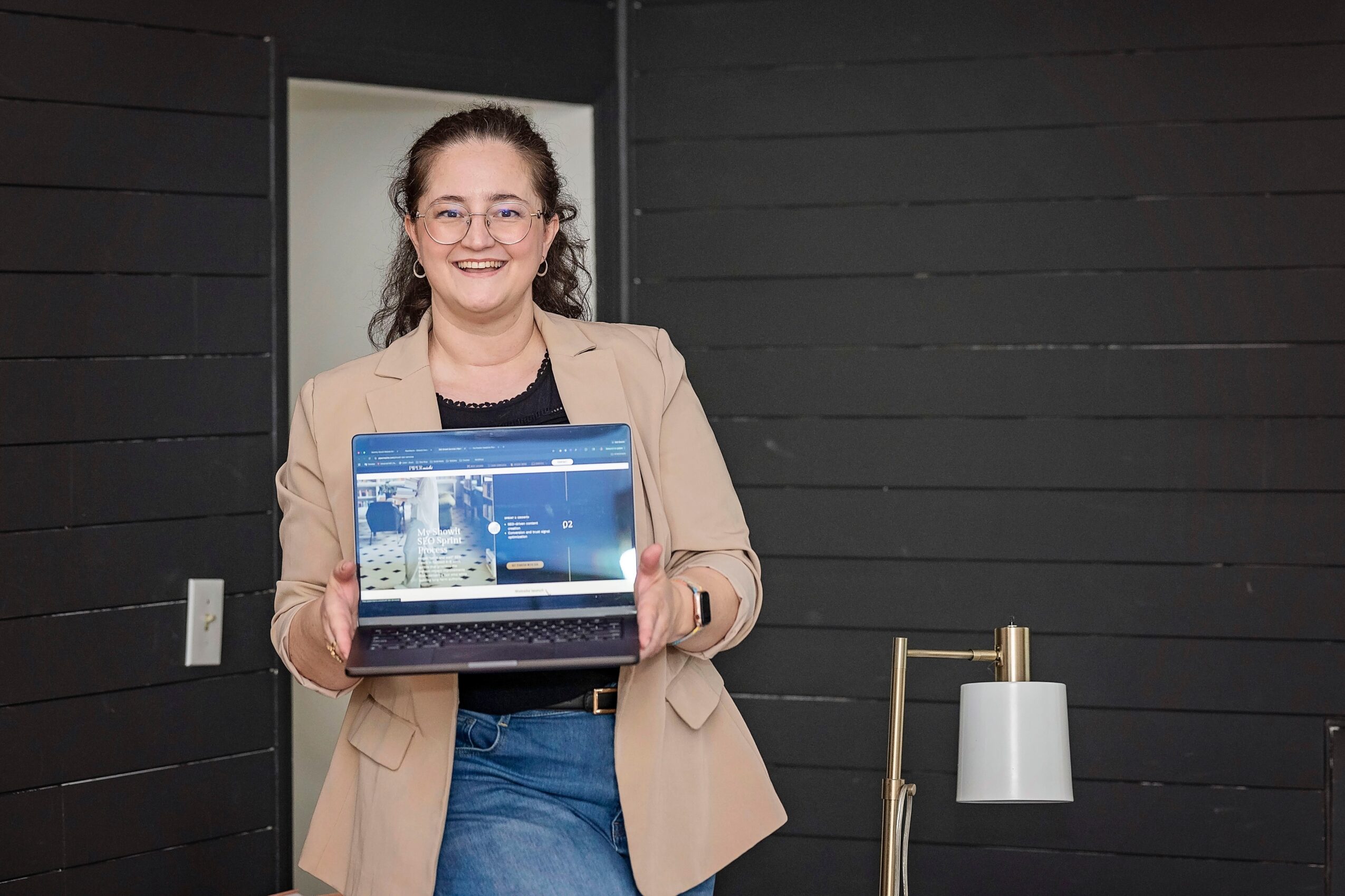 Woman holding a laptop, smiling, standing in front of a dark wall, wearing glasses, a beige blazer, and jeans.