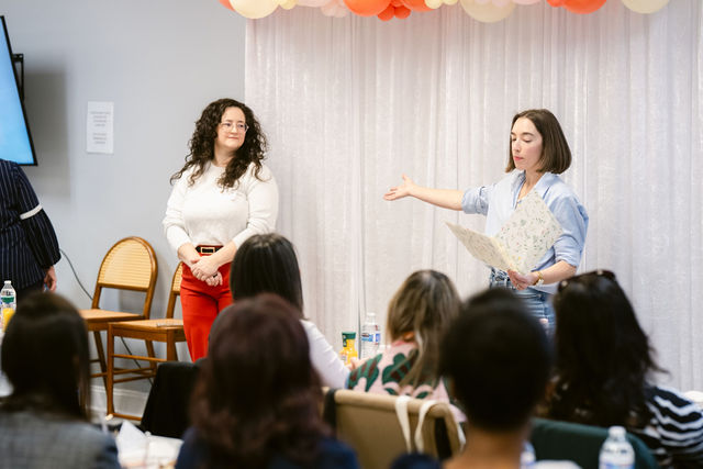 Woman speaking to group in a room with chairs and colorful decorations, audience listening attentively.
