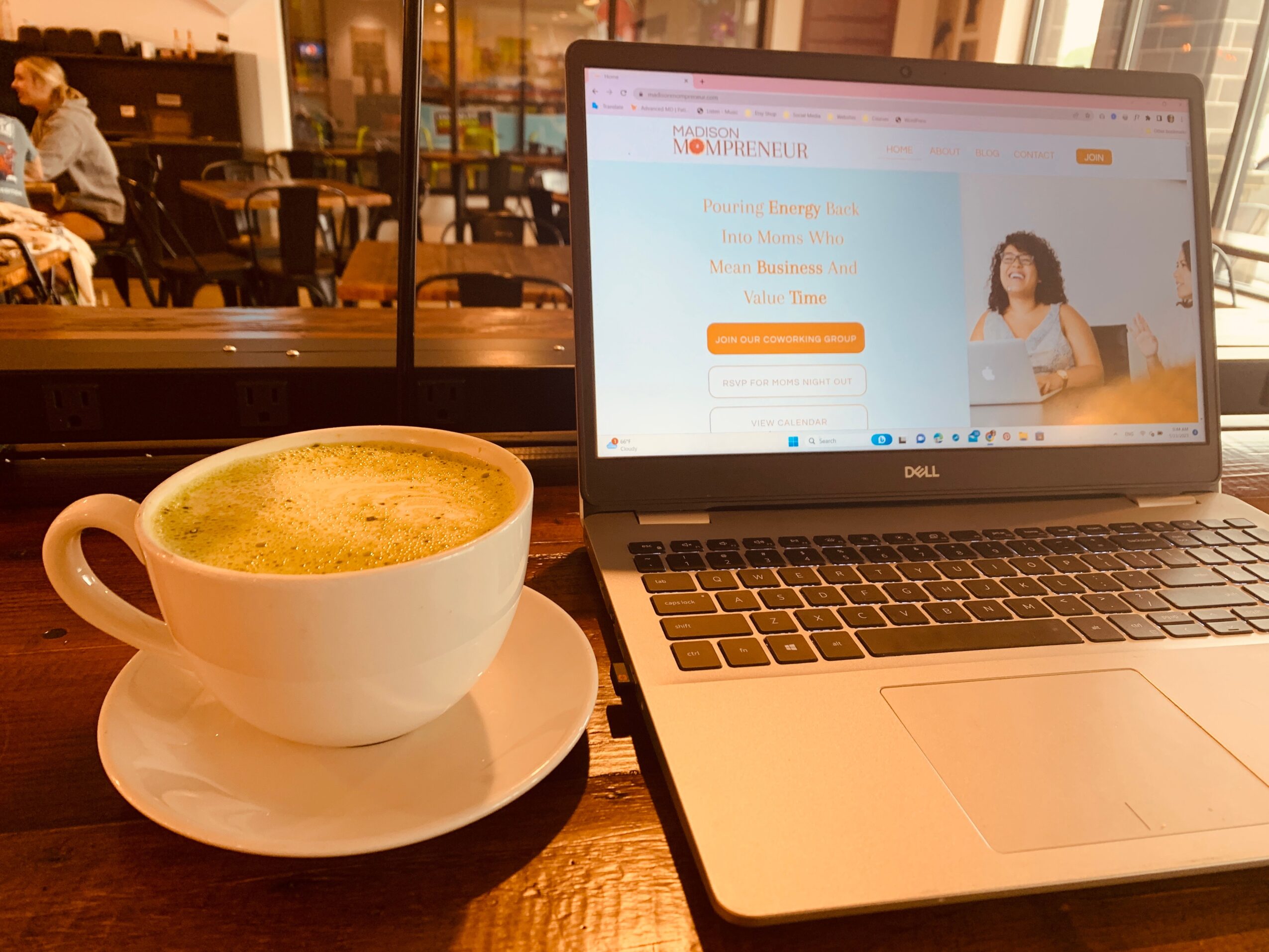 Coffee cup on saucer and open laptop on table in a cafe, with a window and seating area in background.