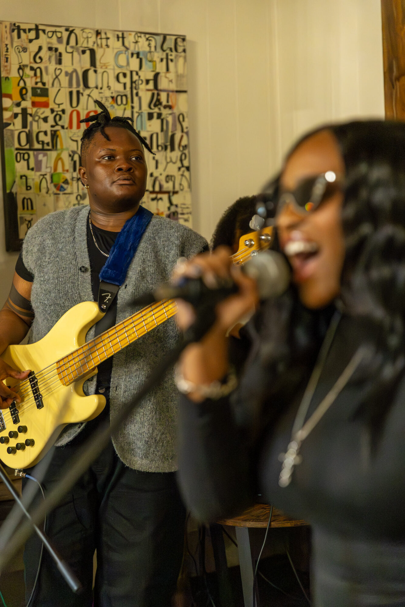 Two women performing music; one singing into a microphone, the other playing a yellow bass guitar, in a room with colorful curtains.