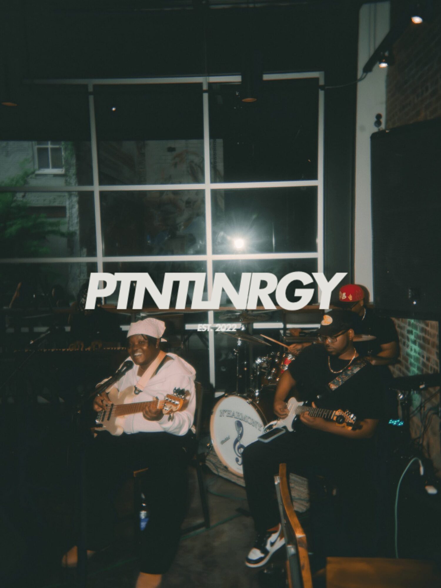 Two musicians playing guitars in a dimly lit indoor space with a large window and a sign reading 'PITNINRGY'.
