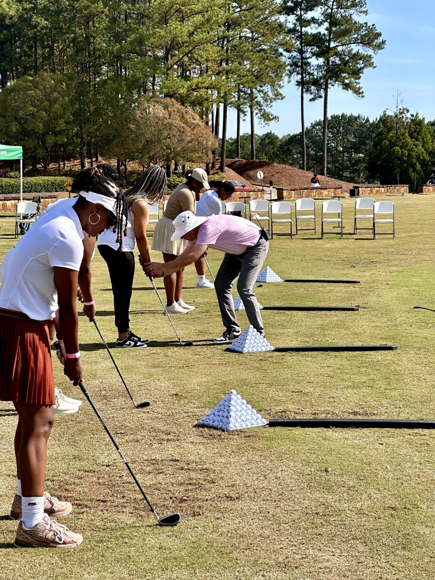 Group of people practicing golf on a grassy field with trees and chairs in the background.