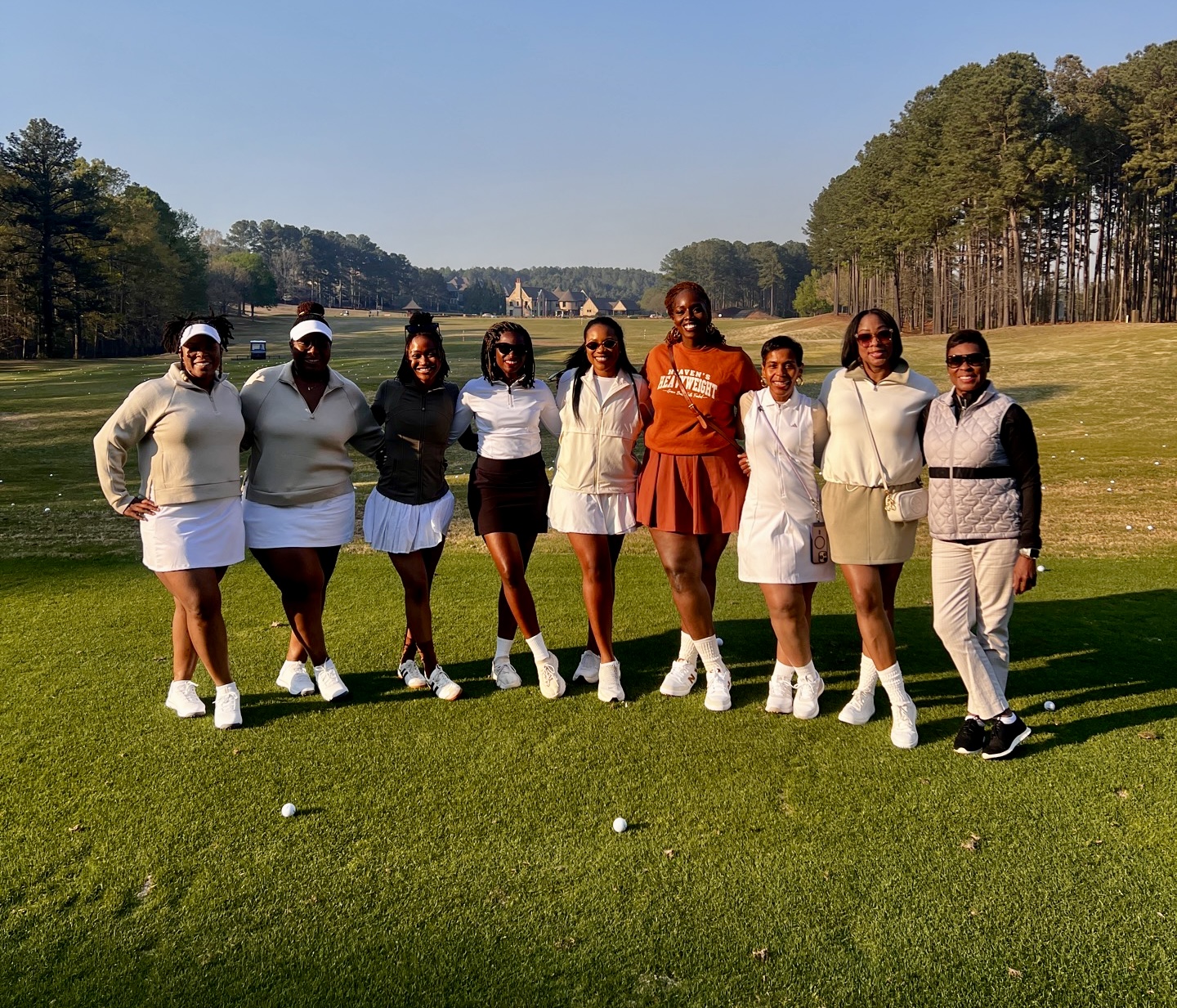 Group of nine women standing on a golf course, dressed in golf attire, with trees and blue sky in background.