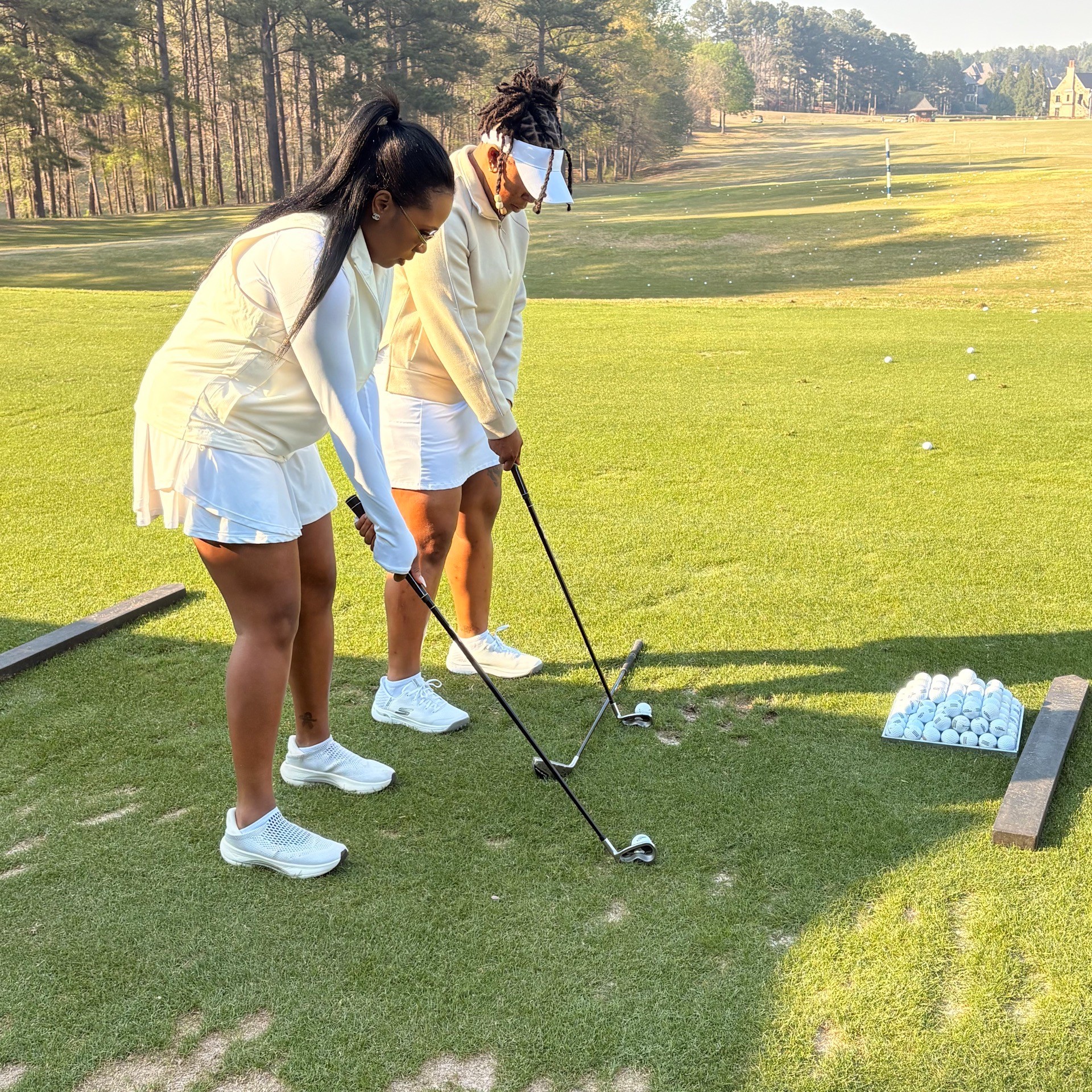 Two young women in white outfits practice golf on a green field, with trees and a clear sky in the background.