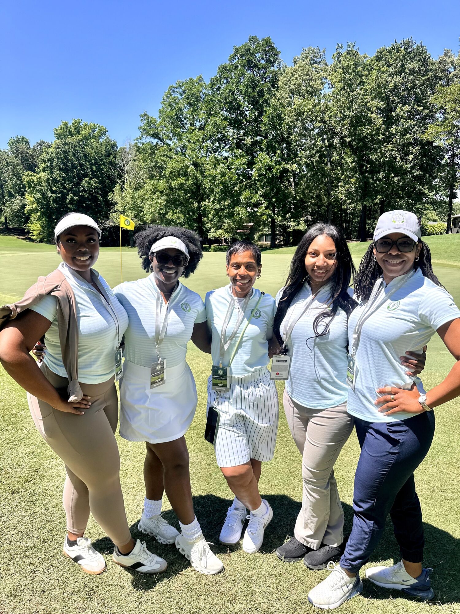 Five women standing outdoors on grass with trees and blue sky in background, smiling and posing for photo.