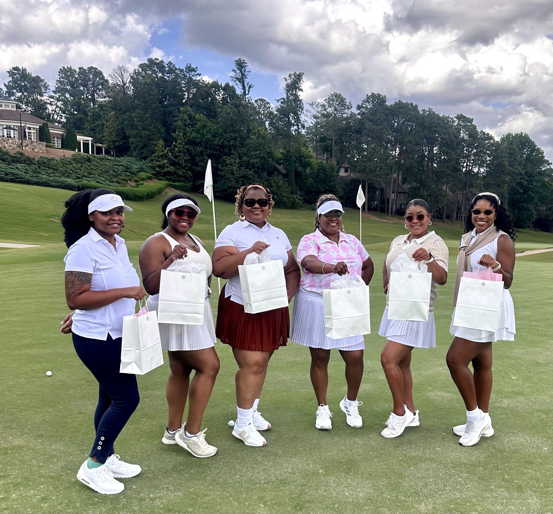 Six women standing on a golf course holding white bags, dressed in white and pink golf attire, with trees and houses in background.