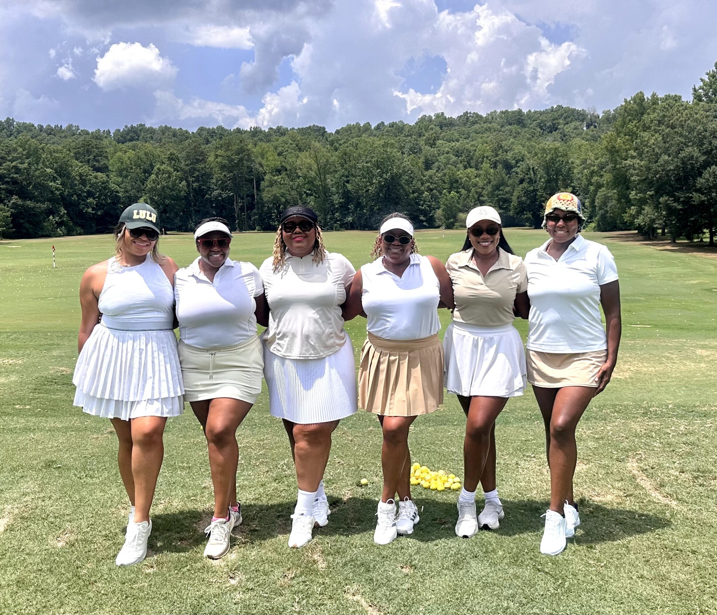 Six women in golf attire standing on a golf course with trees and blue sky in background.