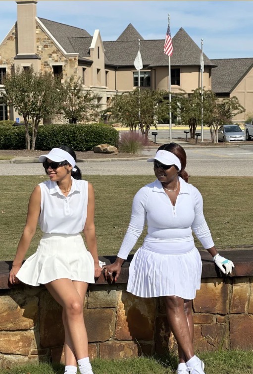 Two women in white outfits and caps sit on a stone wall in a park with buildings and trees behind them.