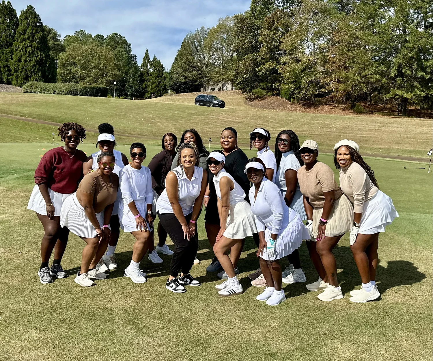 Group of people posing on a golf course with trees and a car in the background, some wearing white outfits and hats.