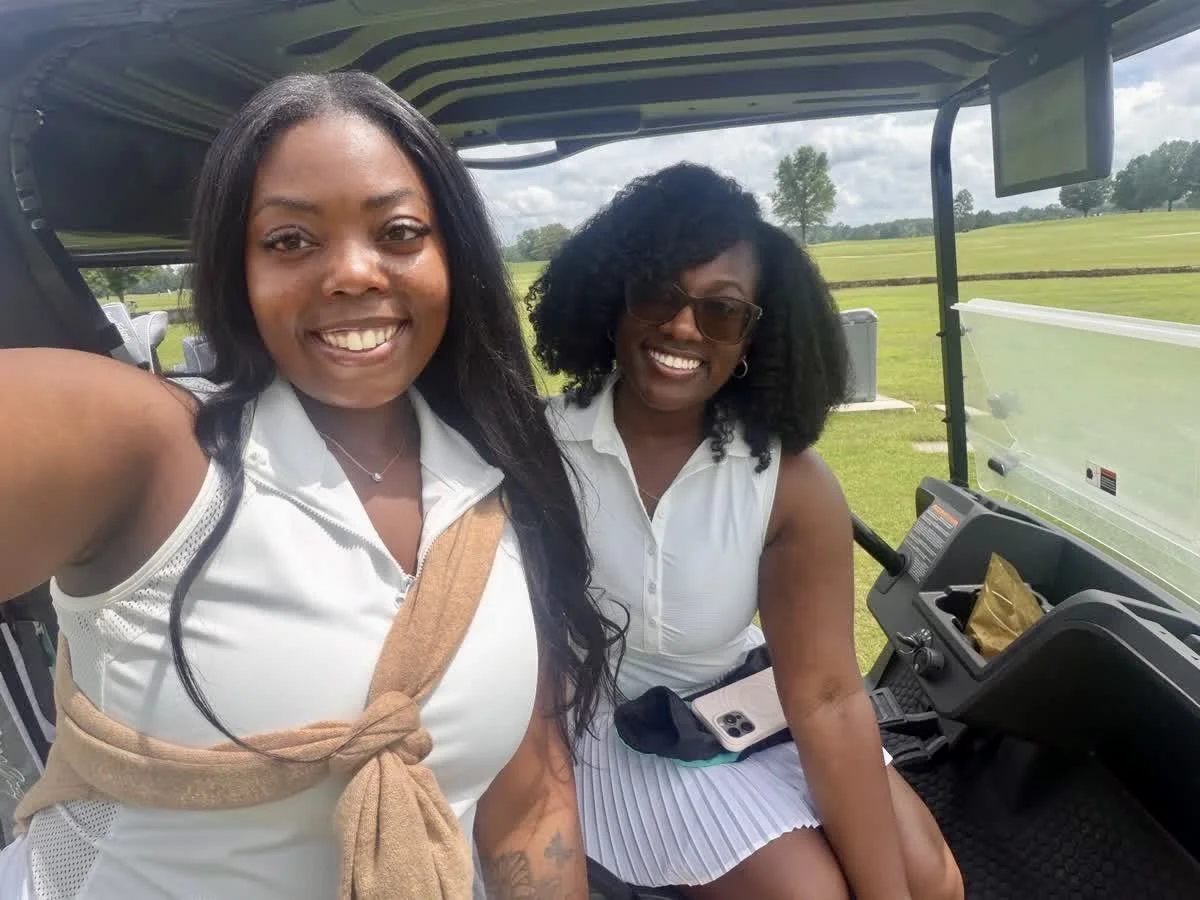 Two women smiling and taking a selfie on a golf cart outdoors with green fields in the background.