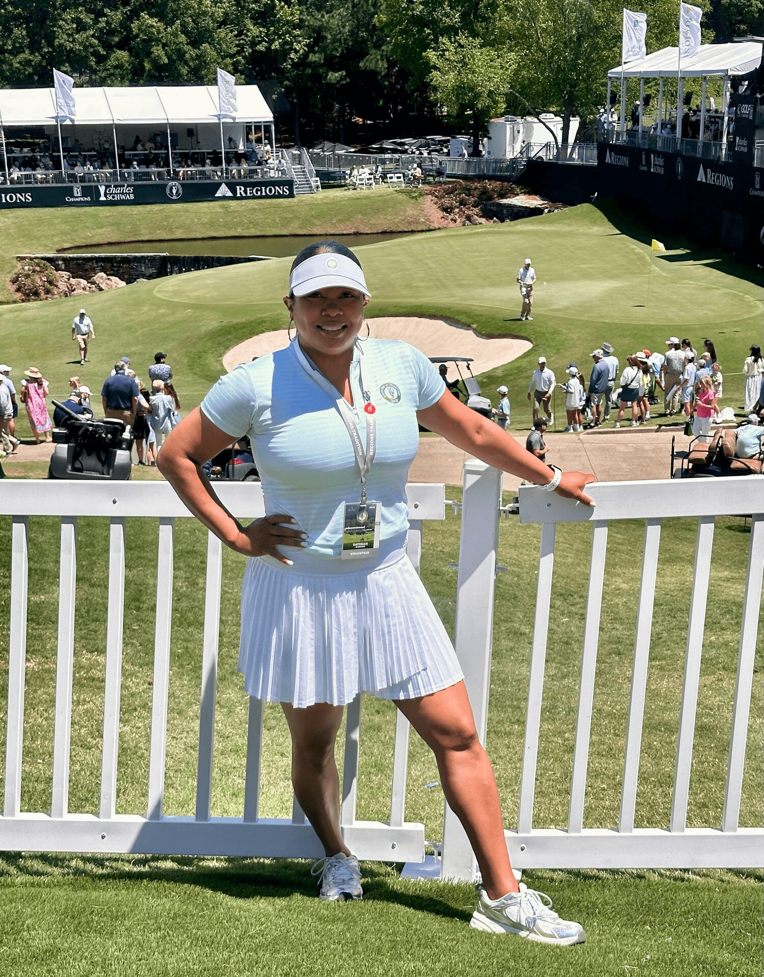 Woman in golf attire standing by a white fence at a golf course, with spectators and a green in the background.