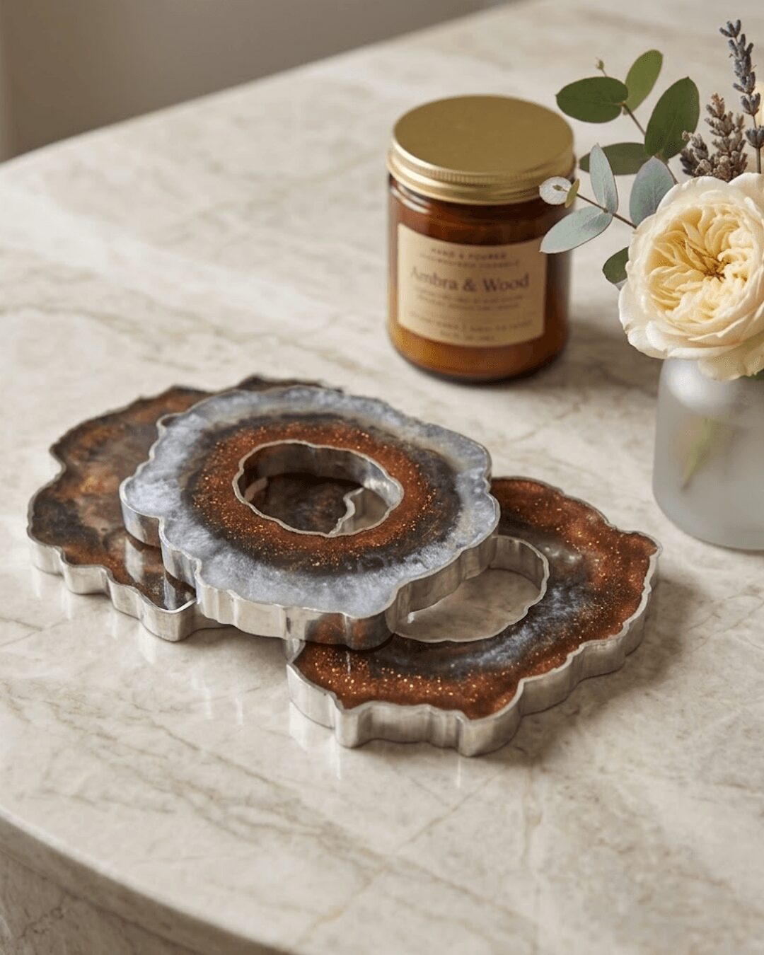 Three agate coasters stacked on a marble surface with a jar of honey and a flower arrangement in a white vase nearby.