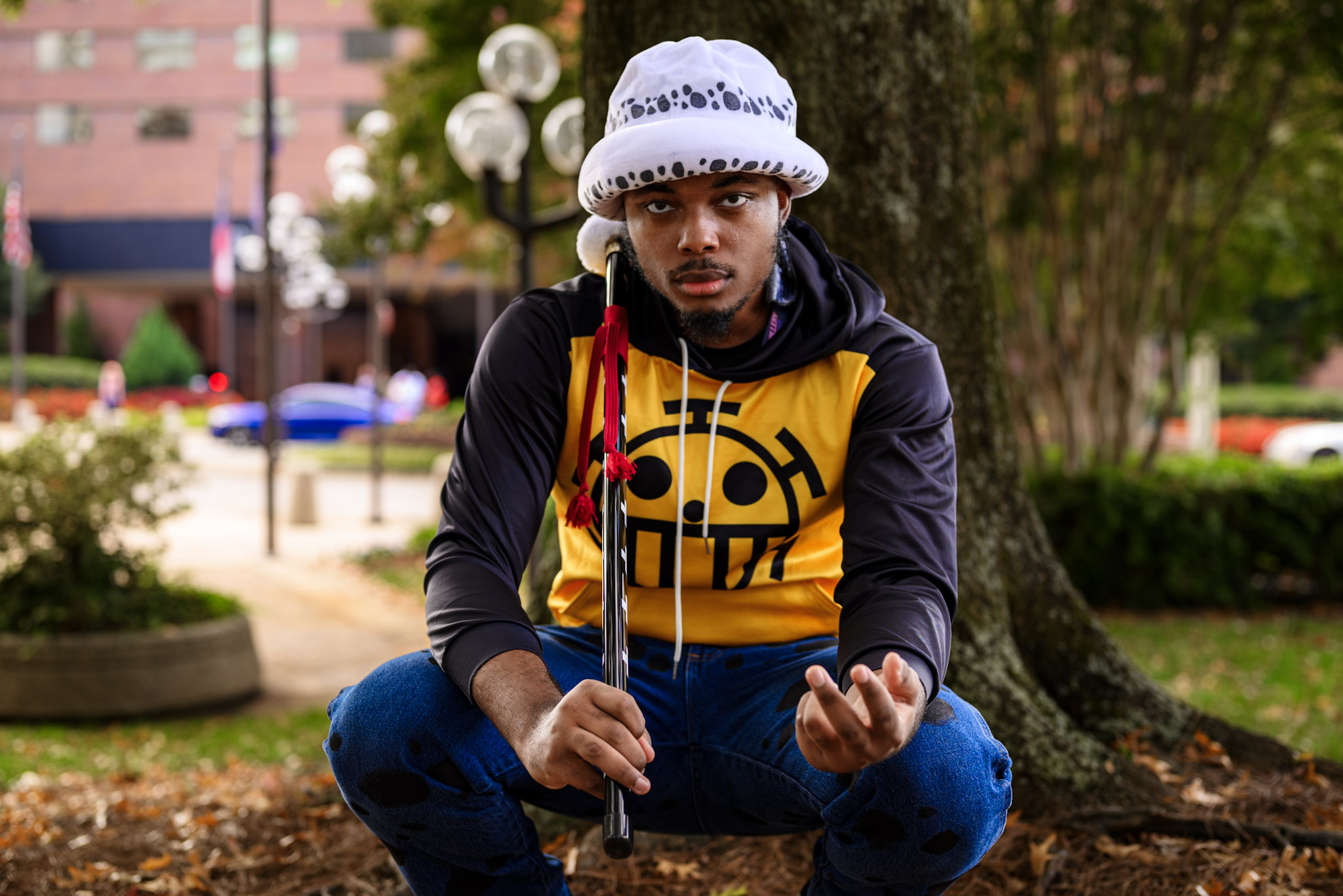 Young man sitting outdoors near a tree, wearing a white hat, yellow and black hoodie, and blue pants, holding a stick.