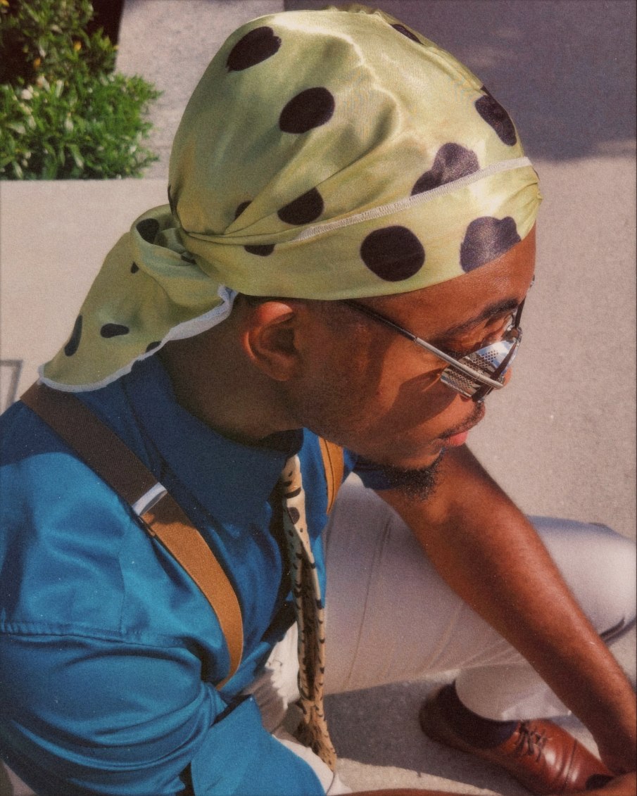 Person wearing a polka dot headscarf, sunglasses, blue jacket, sitting outdoors on a sunny day.