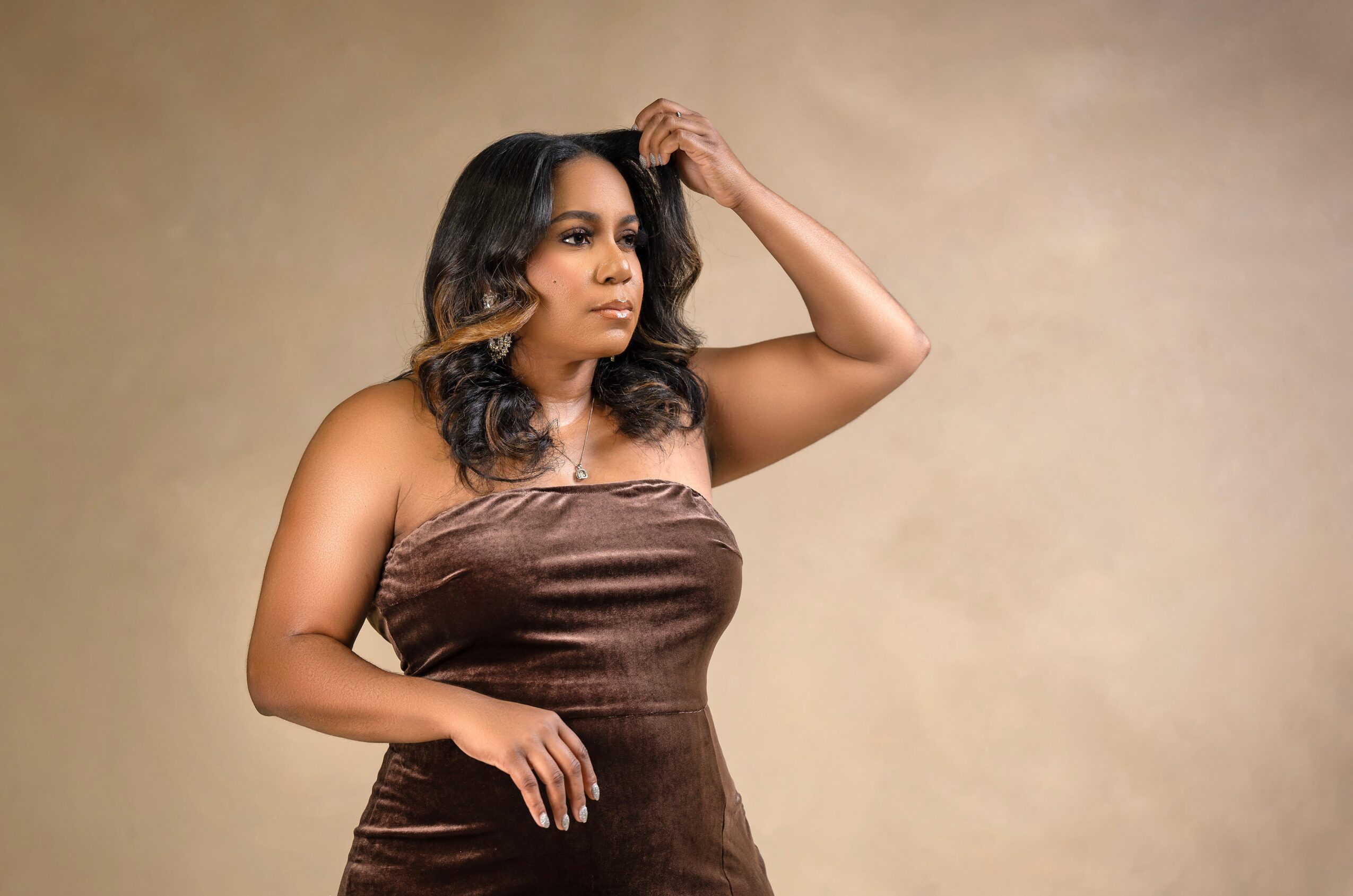 Woman with dark hair and wavy curls, wearing a strapless brown dress, posing with her hand on her head against a beige background.