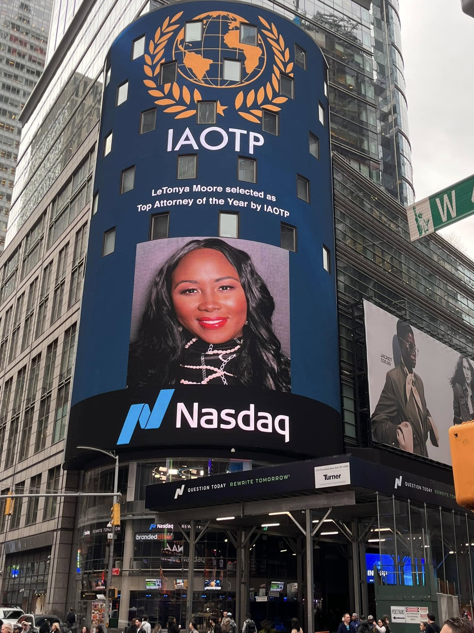 Digital billboard displaying a woman's portrait, Nasdaq logo, and IAOTP emblem in an urban setting with tall buildings.