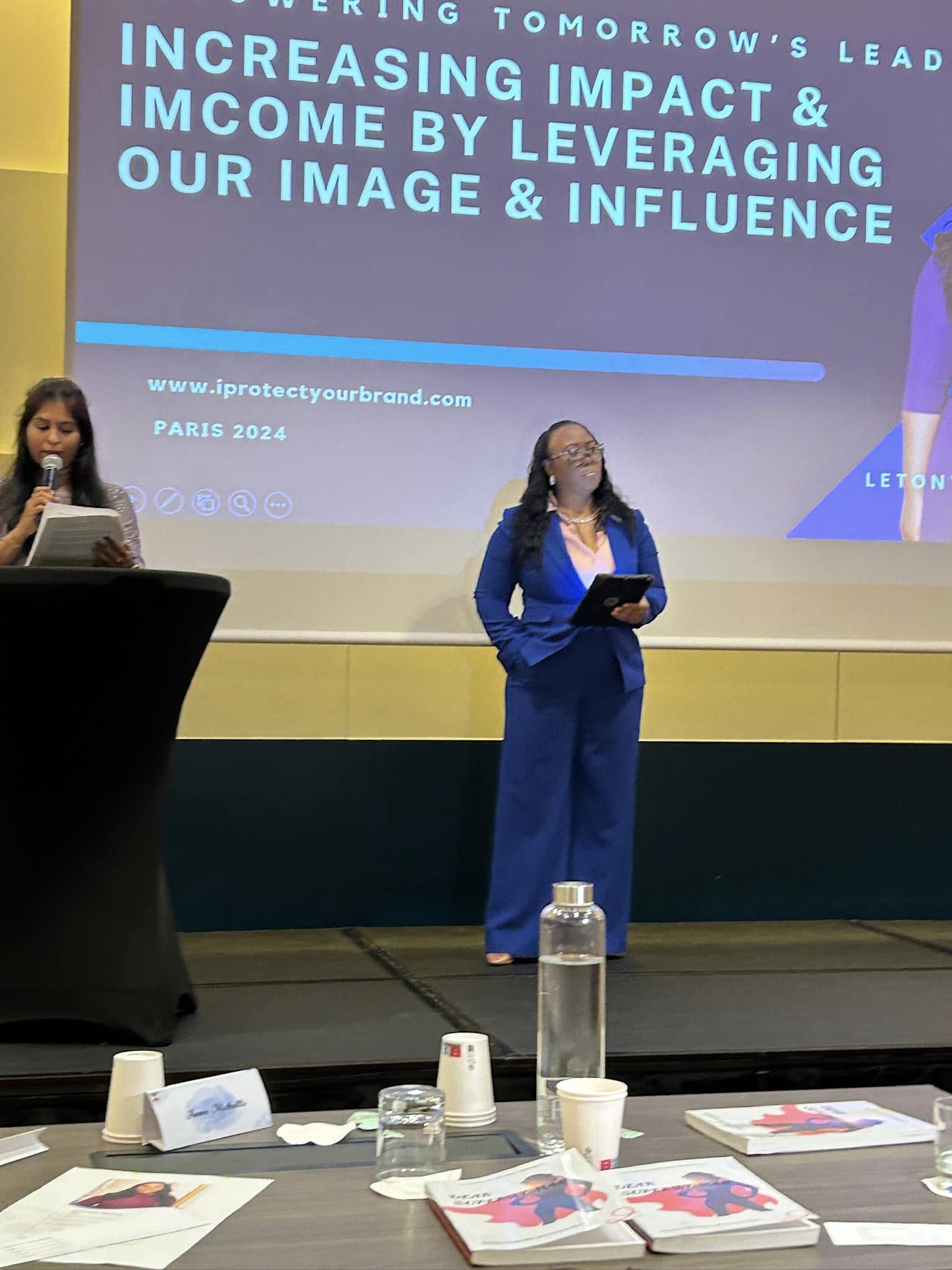 Two women stand on stage in front of a large screen, one holding a tablet, with a table and water bottles in front.