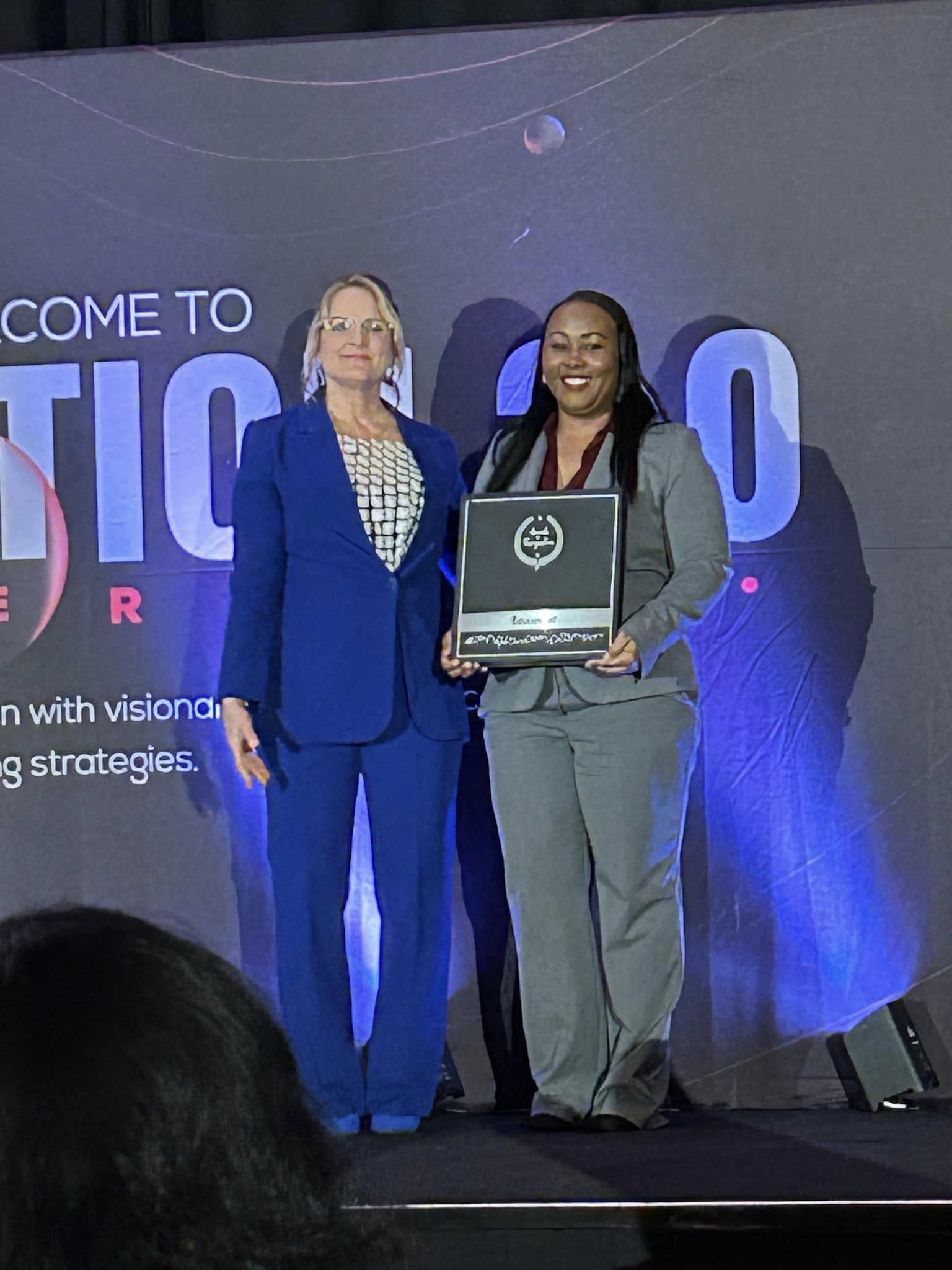 Two women standing on stage, one holding a framed award, with a large screen behind them displaying text and graphics.
