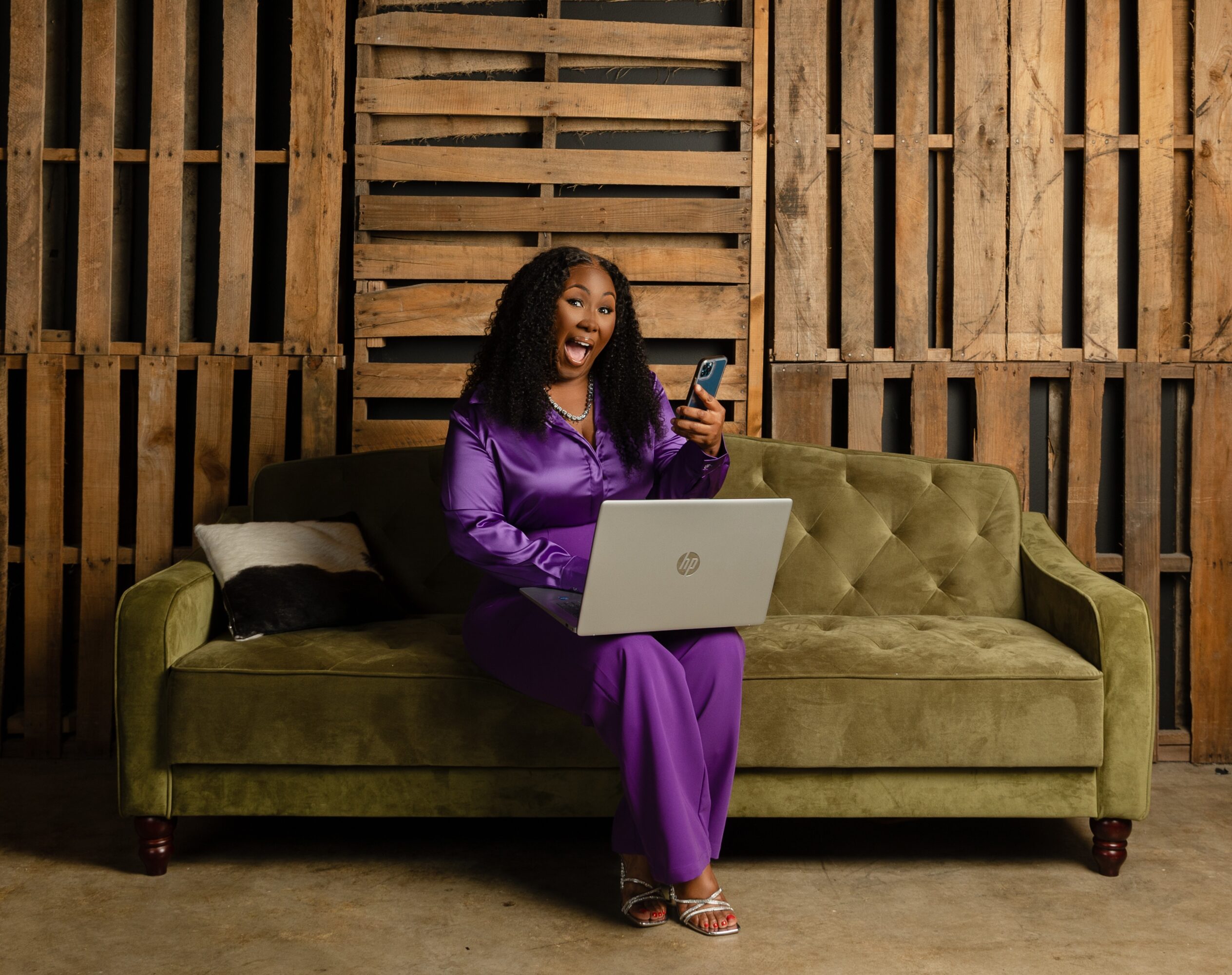 Woman in purple outfit sitting on a green sofa, holding a phone and laptop, smiling, in a wooden-paneled room.