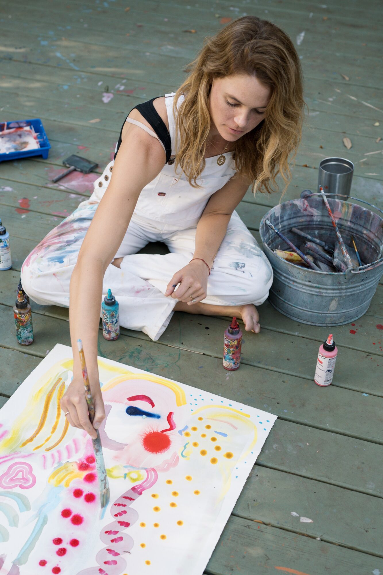 Young woman sitting on floor painting colorful dots and shapes on large paper, surrounded by art supplies.