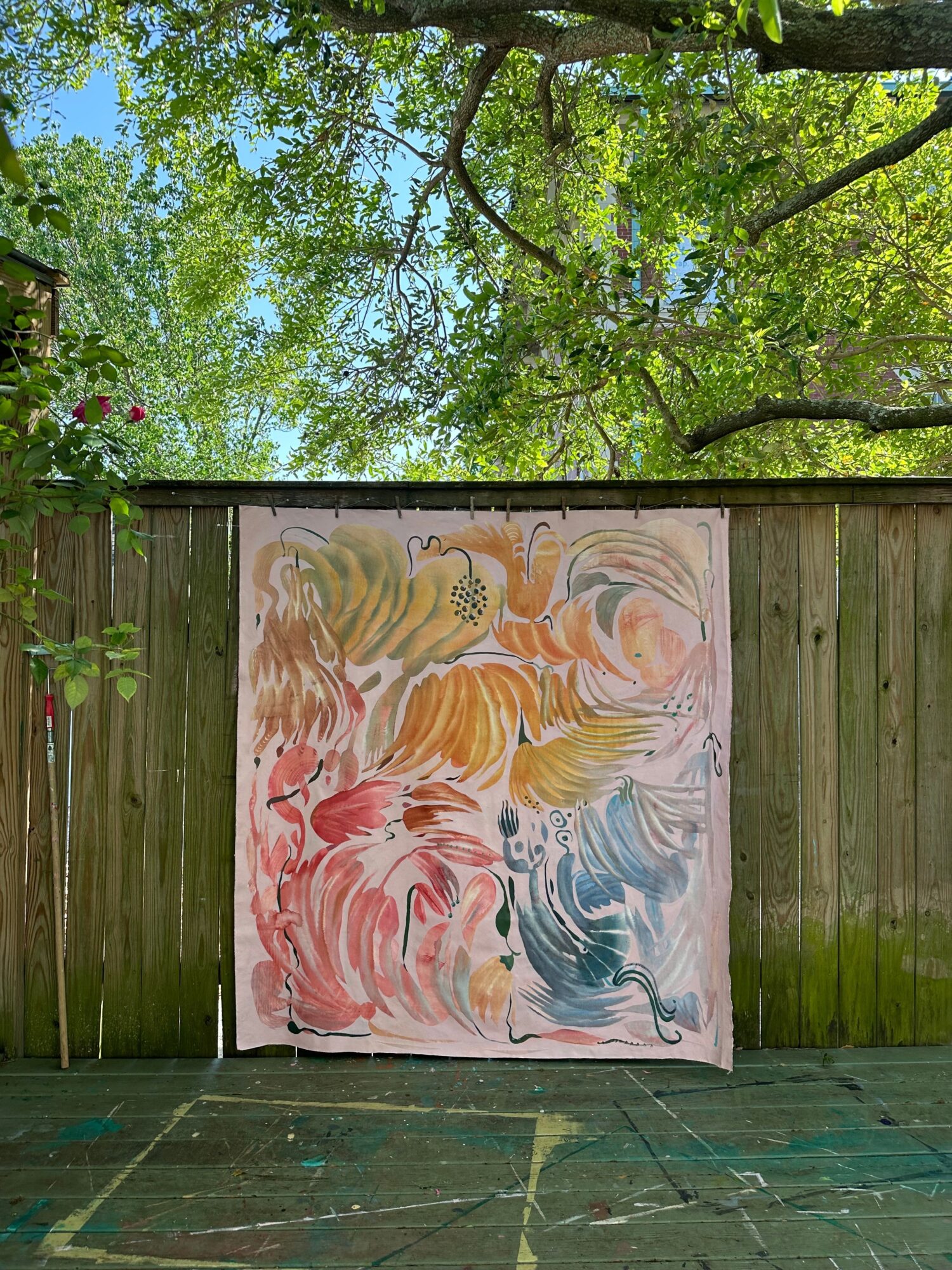 Colorful abstract painting on a white canvas, outdoors against a wooden fence, with green trees and sky in background.