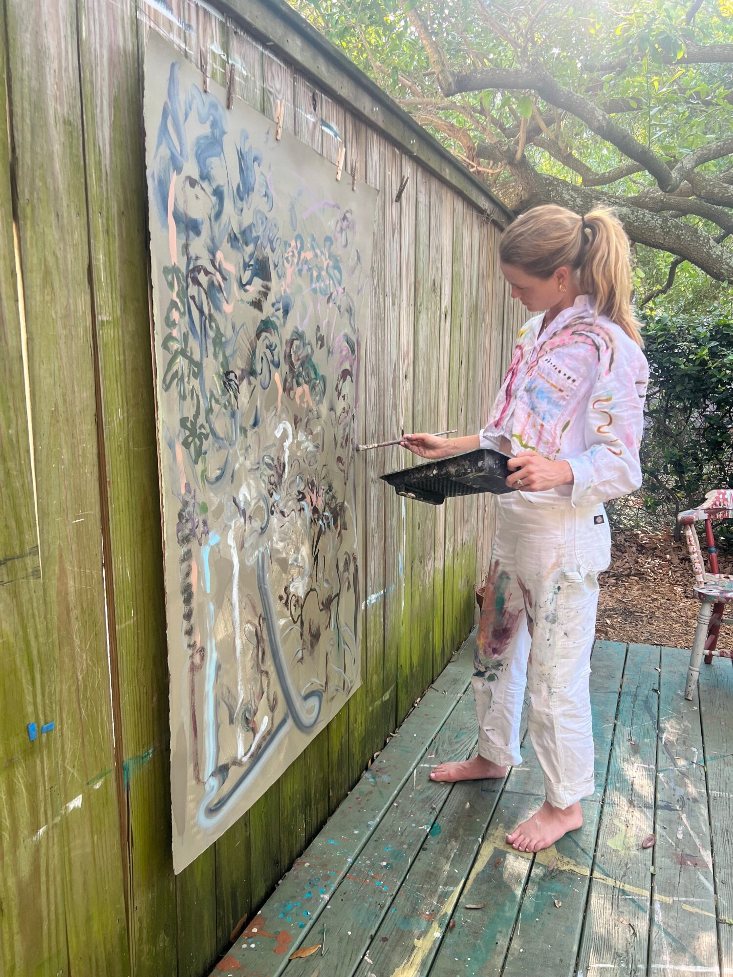 Woman with blonde hair in a pink and white striped shirt and white pants stands on a wooden deck, holding a phone, next to a large abstract painting.