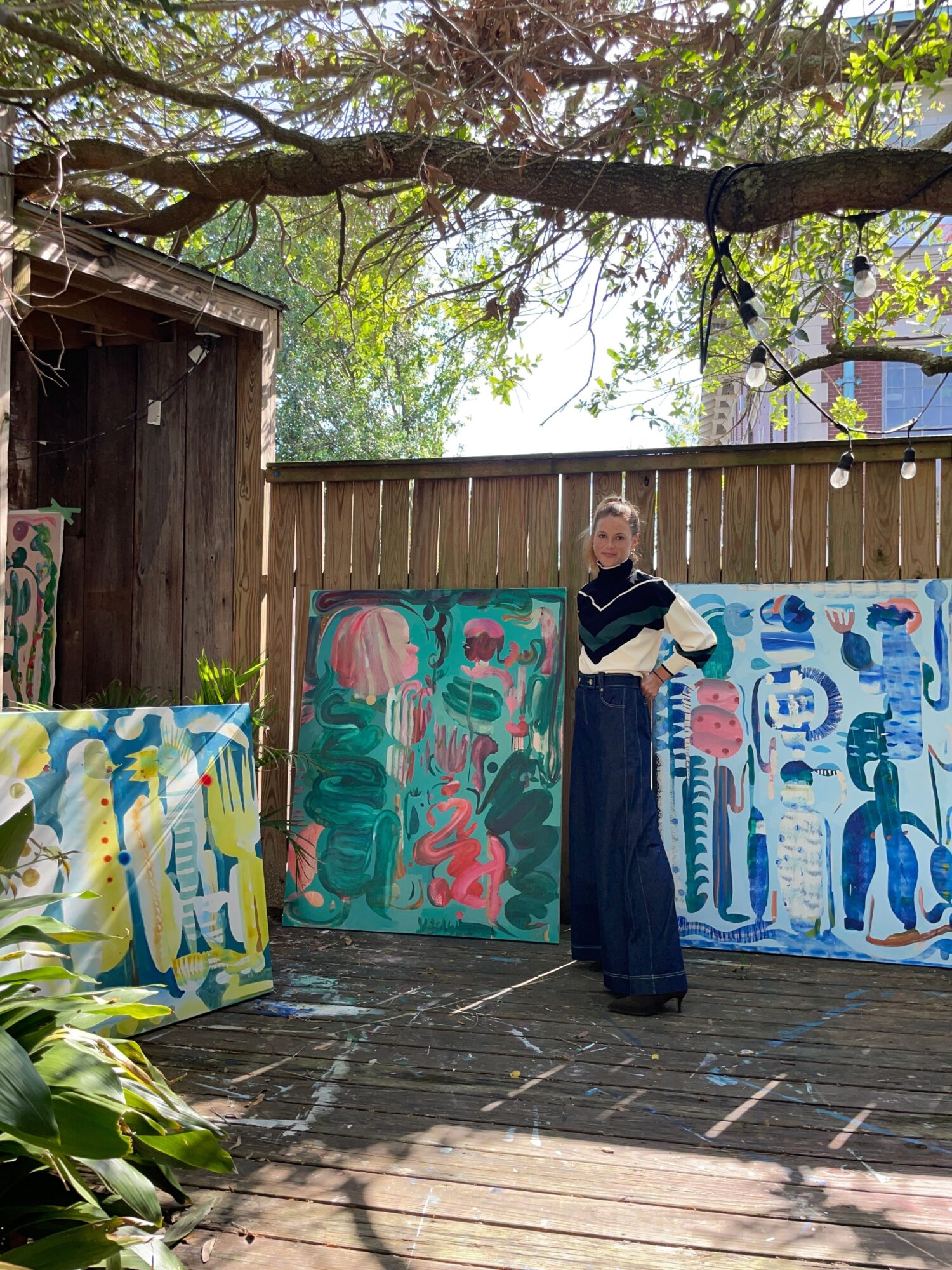 Person standing outdoors near colorful paintings on display, trees and wooden fence in background.