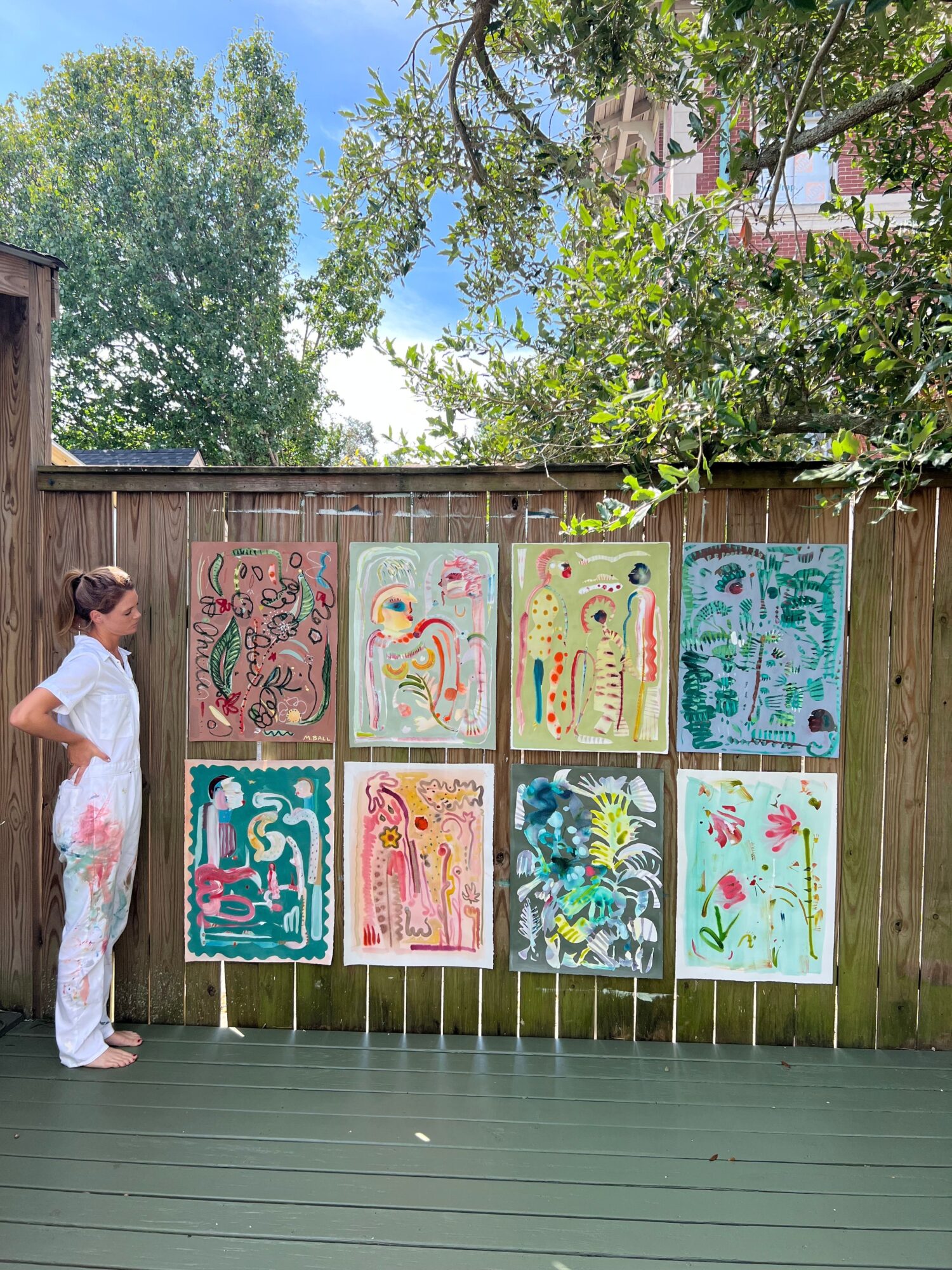 Child in white dress with pink floral pattern standing outdoors near colorful paintings on a wooden fence, trees and sky in background.