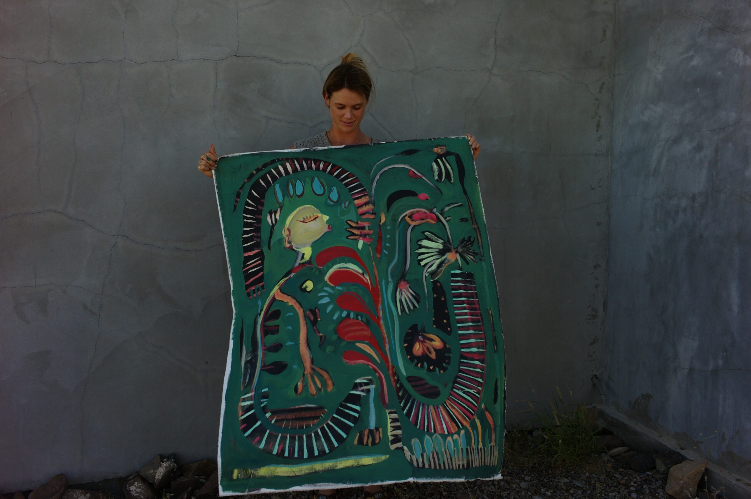 Person holding a colorful, abstract painting with a green background and various shapes, against a plain wall.