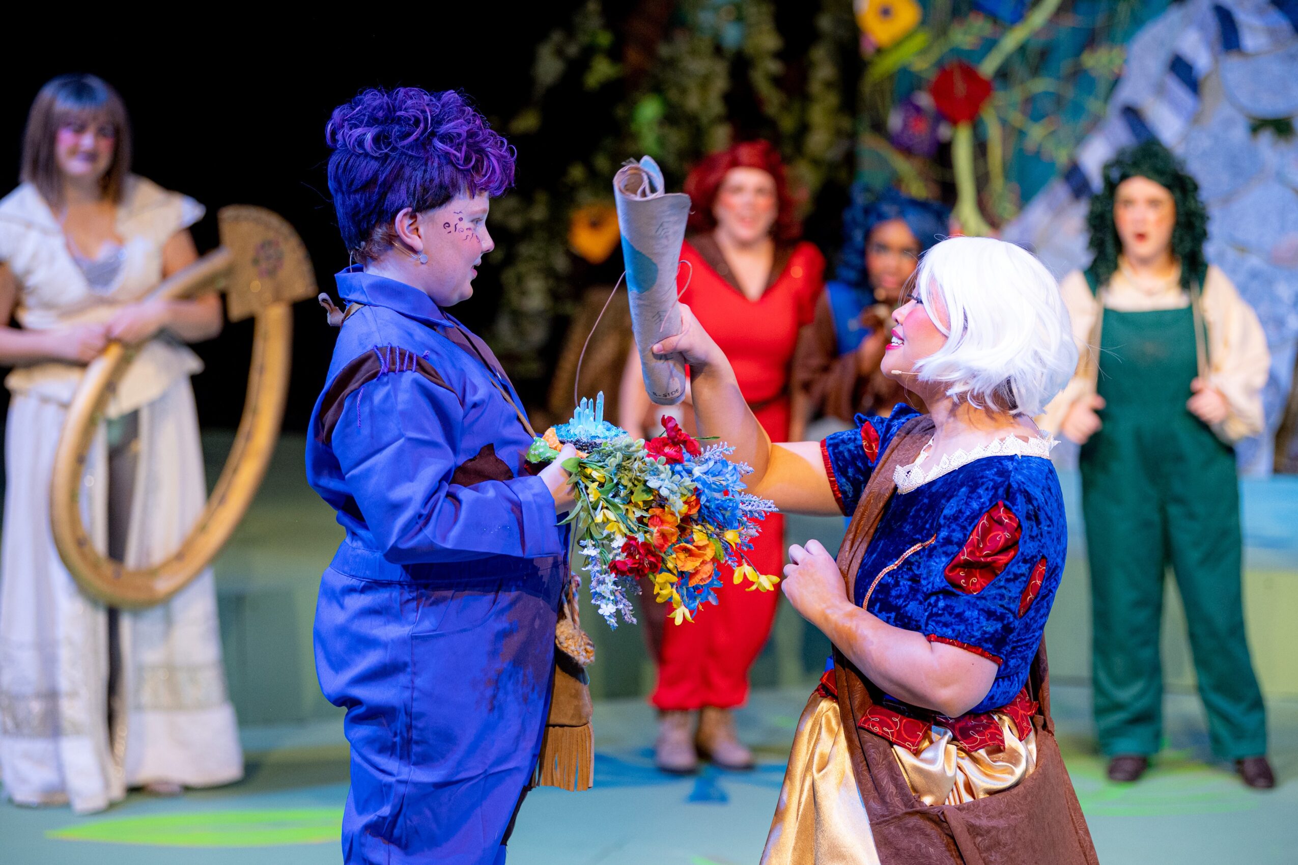 Person with white hair and blue dress hands a bouquet to a person with purple hair in a blue outfit, stage background.