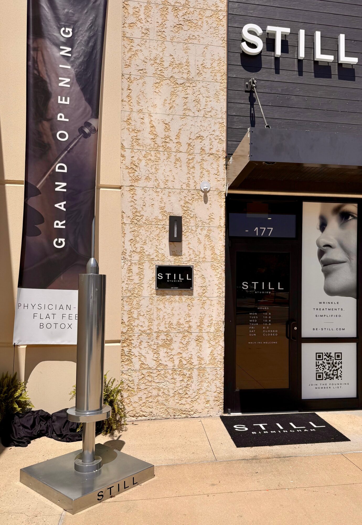 Store entrance with signage, a poster of a woman's face, and a QR code, outside a building with textured wall.