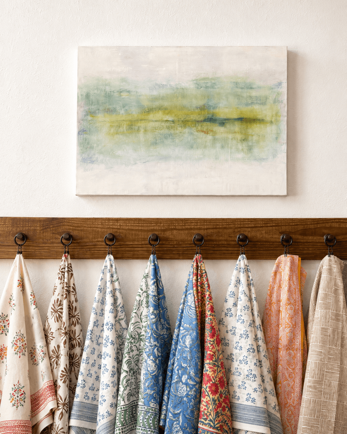 Watercolor landscape painting hangs above a wooden coat rack with various fabric swatches. The wall is light-colored.