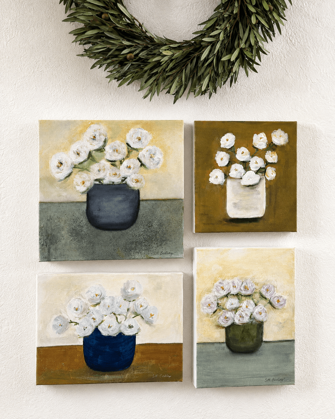 Four small paintings of flower vases arranged in a grid, with a green wreath above, on a white surface.