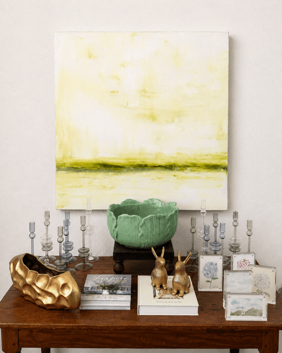 Decorative table with a large green bowl, gold sculpture, two rabbit figurines, and framed pictures, against a white wall with abstract painting.