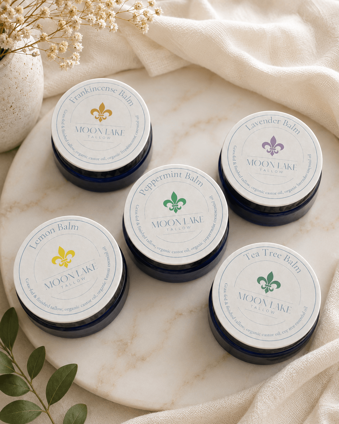 Five small jars with labels arranged on a marble surface, with a cloth and green leaves nearby.