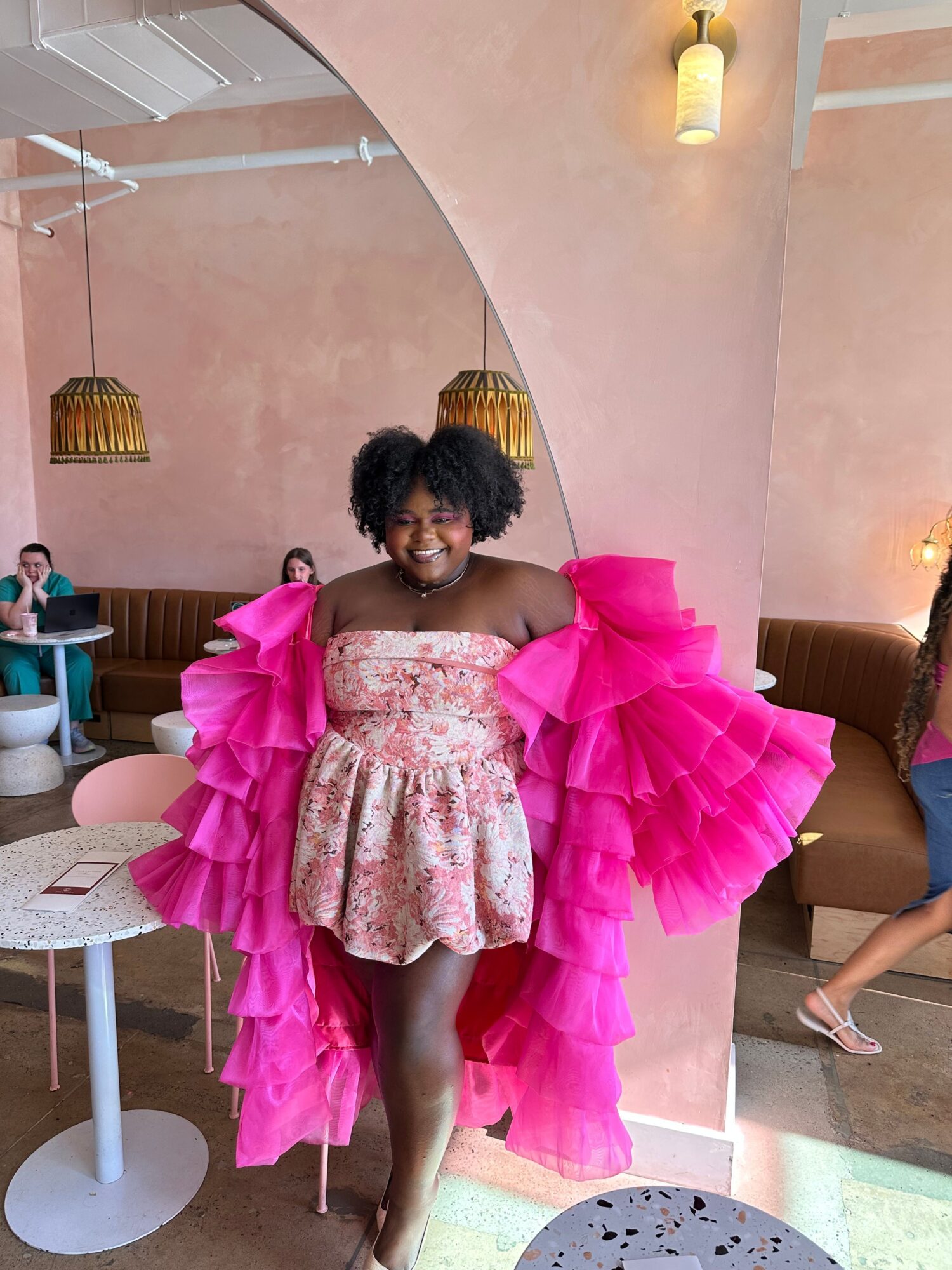 Child in pink ruffled dress standing indoors near a pink wall with a white table and a person in the background.
