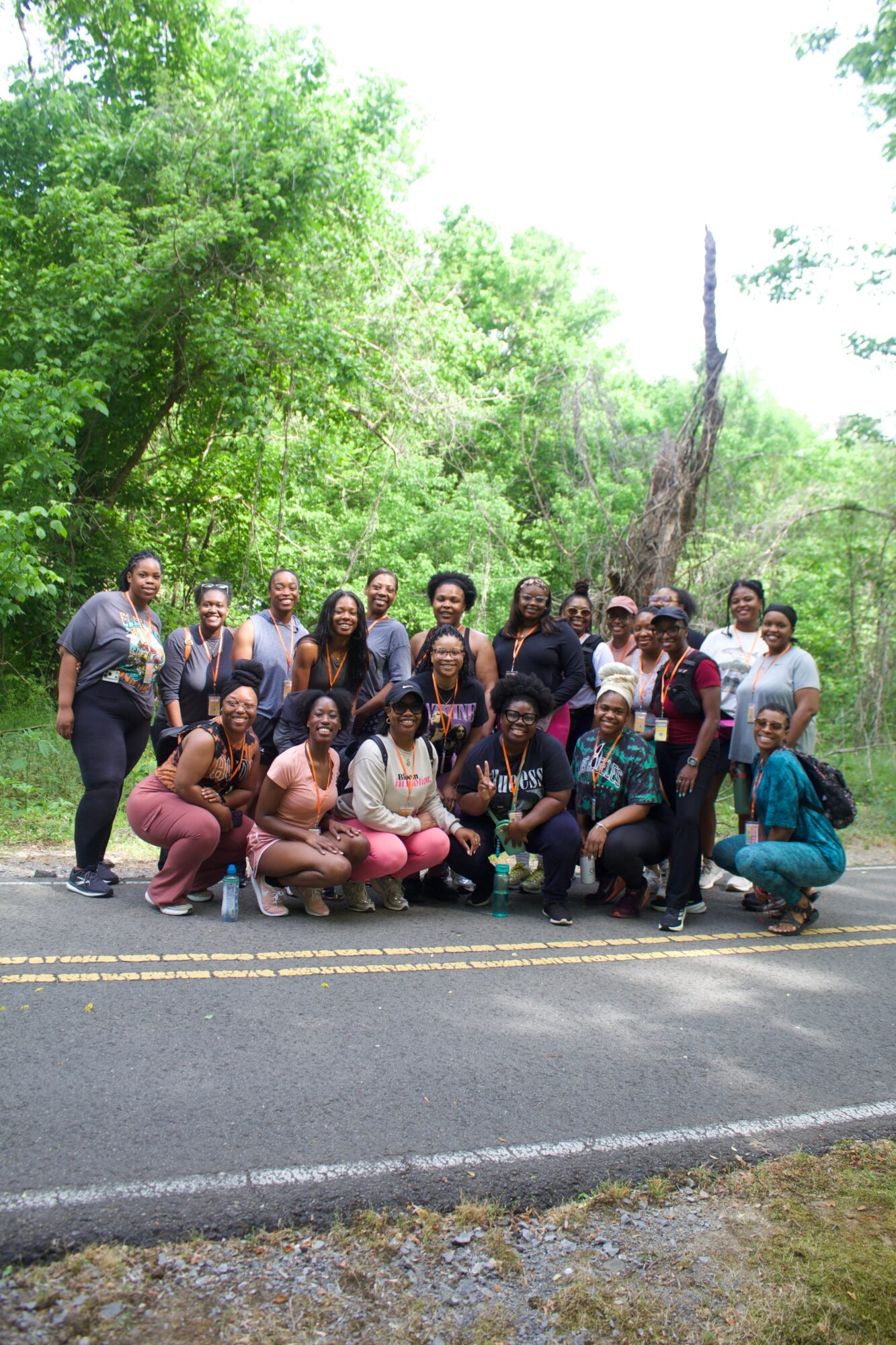 Group of people outdoors on a road with green trees in the background, some standing and some crouching or sitting.
