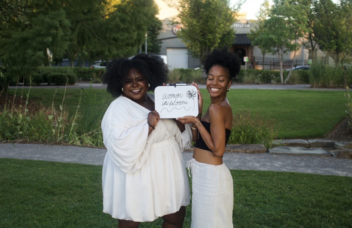 Two women smiling outdoors, holding a sign with handwritten text, standing on grass with trees and a pathway behind them.
