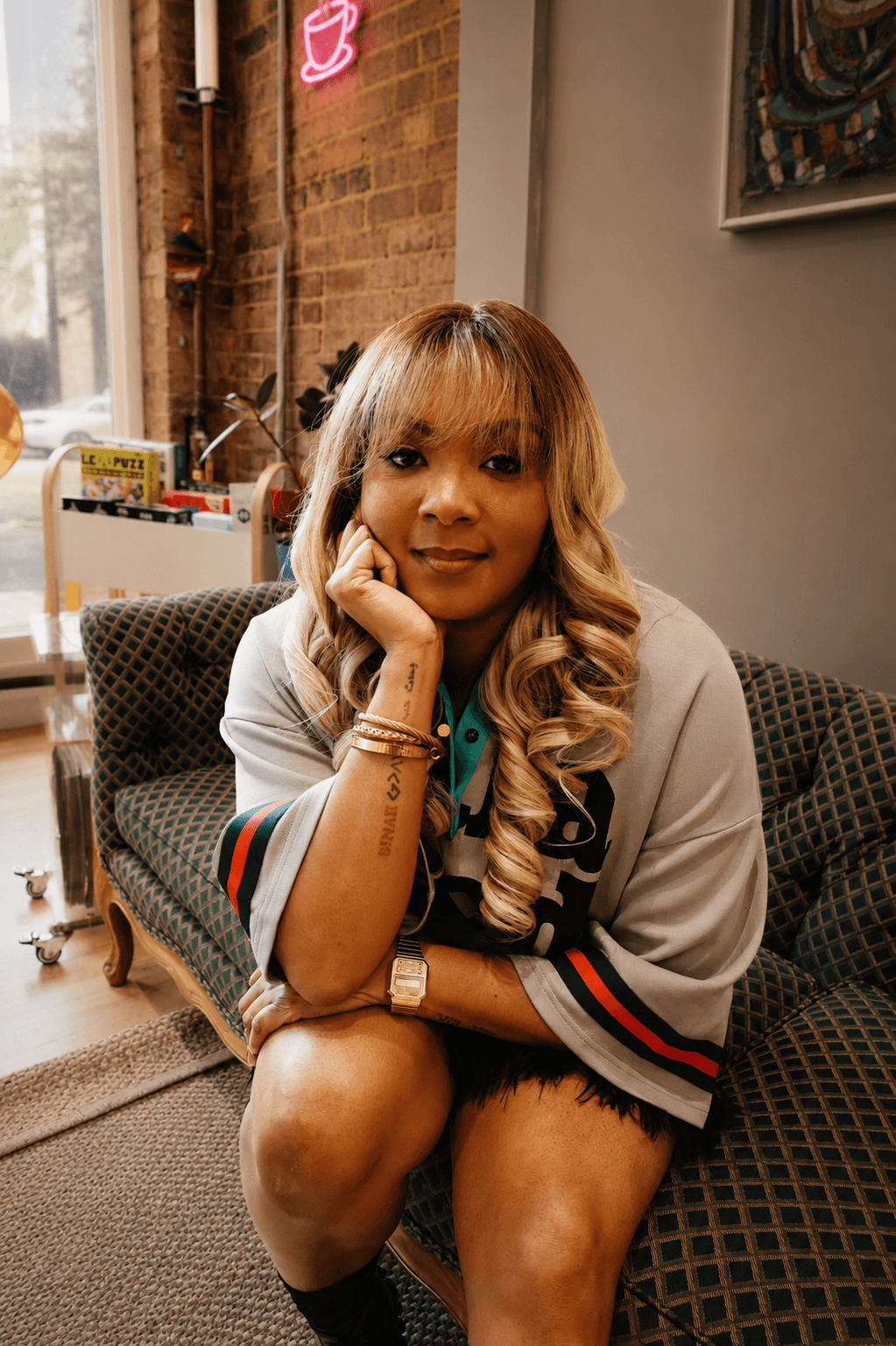 Woman with long wavy hair sitting on a patterned sofa in a room with brick wall and window.