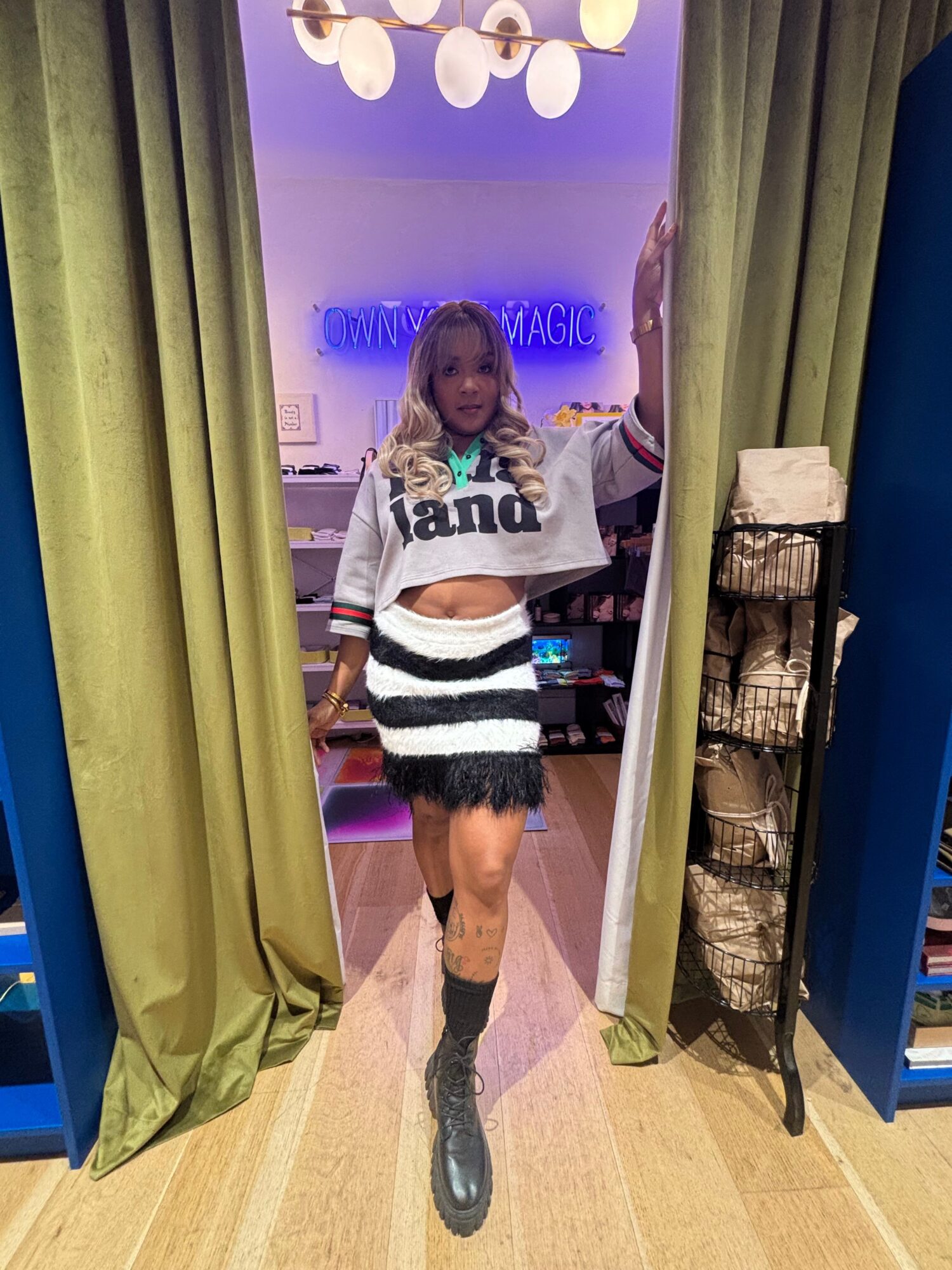 Person standing in a room with purple and green curtains, wearing a striped sweater and black boots.
