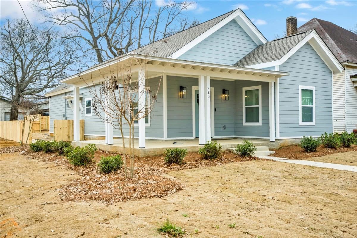Blue house with a covered porch, surrounded by a yard with small bushes and a tree, under a clear sky.