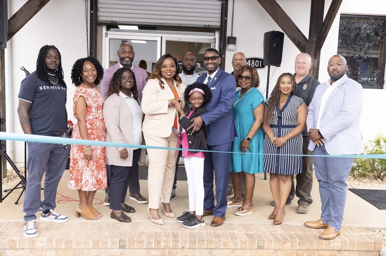 Group of people standing outside a building, some holding a ribbon for a ceremony, smiling for the photo.