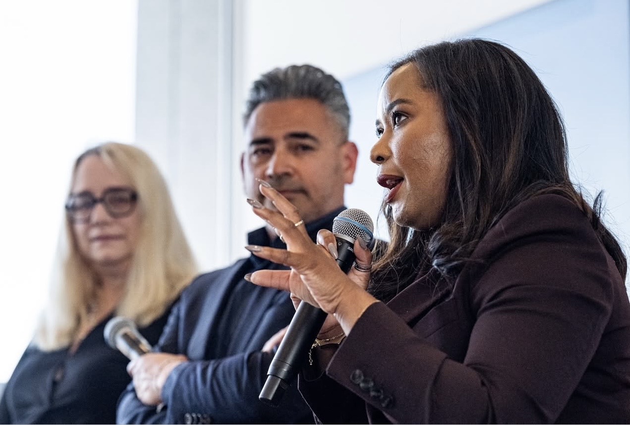 Three people sitting, woman speaking into a microphone, others listening, in a bright room.